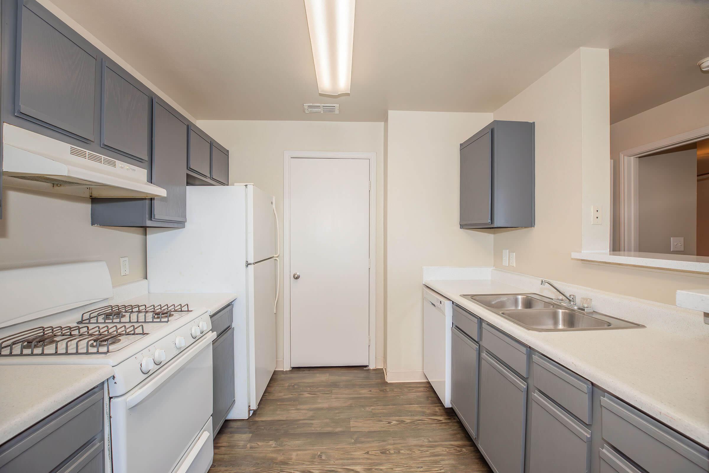 A modern kitchen with grey cabinets, white appliances, and a spacious countertop. Features a gas stove, refrigerator, and double sink. The walls are light-colored, and the flooring is a warm wood tone. A door leads to another room, and a window provides natural light.