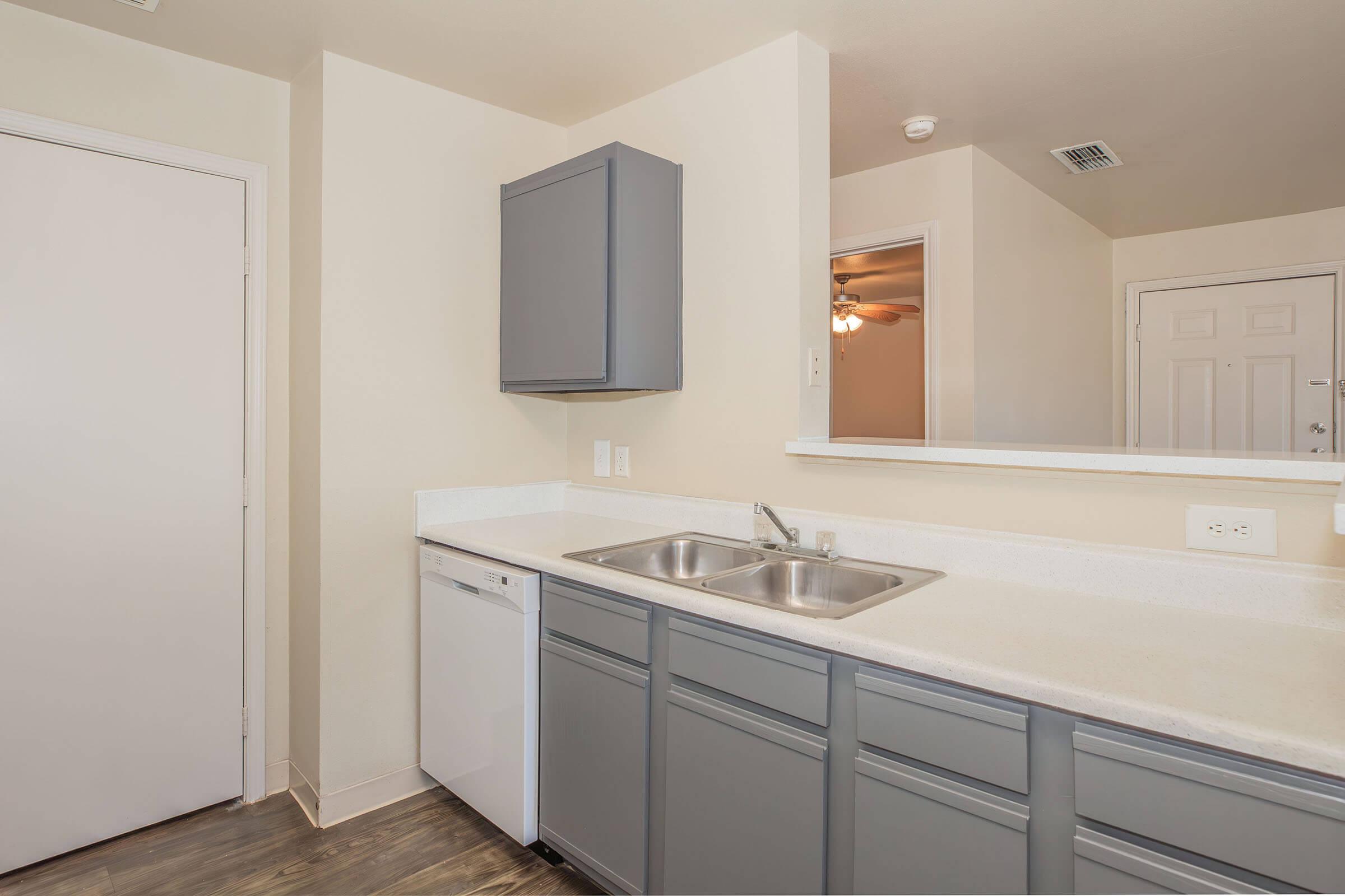 A modern kitchen featuring gray cabinets, a double sink with a chrome faucet, and a dishwasher. The walls are painted in a light color, and there's a doorway leading to another room. A mirror reflects part of the space, enhancing the openness of the kitchen area.