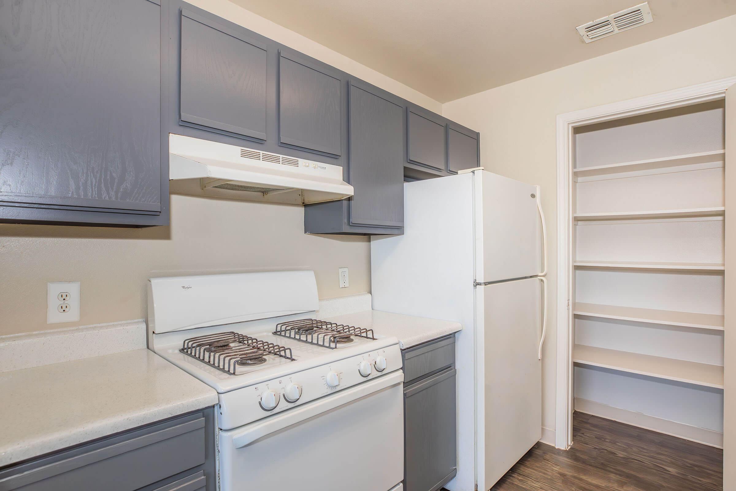 A modern kitchen featuring gray cabinets, a white gas stove, and a white refrigerator. The countertop is light-colored, and there is an exhaust hood above the stove. A spacious pantry with shelves is visible in the background, providing additional storage space. The flooring is dark wood.