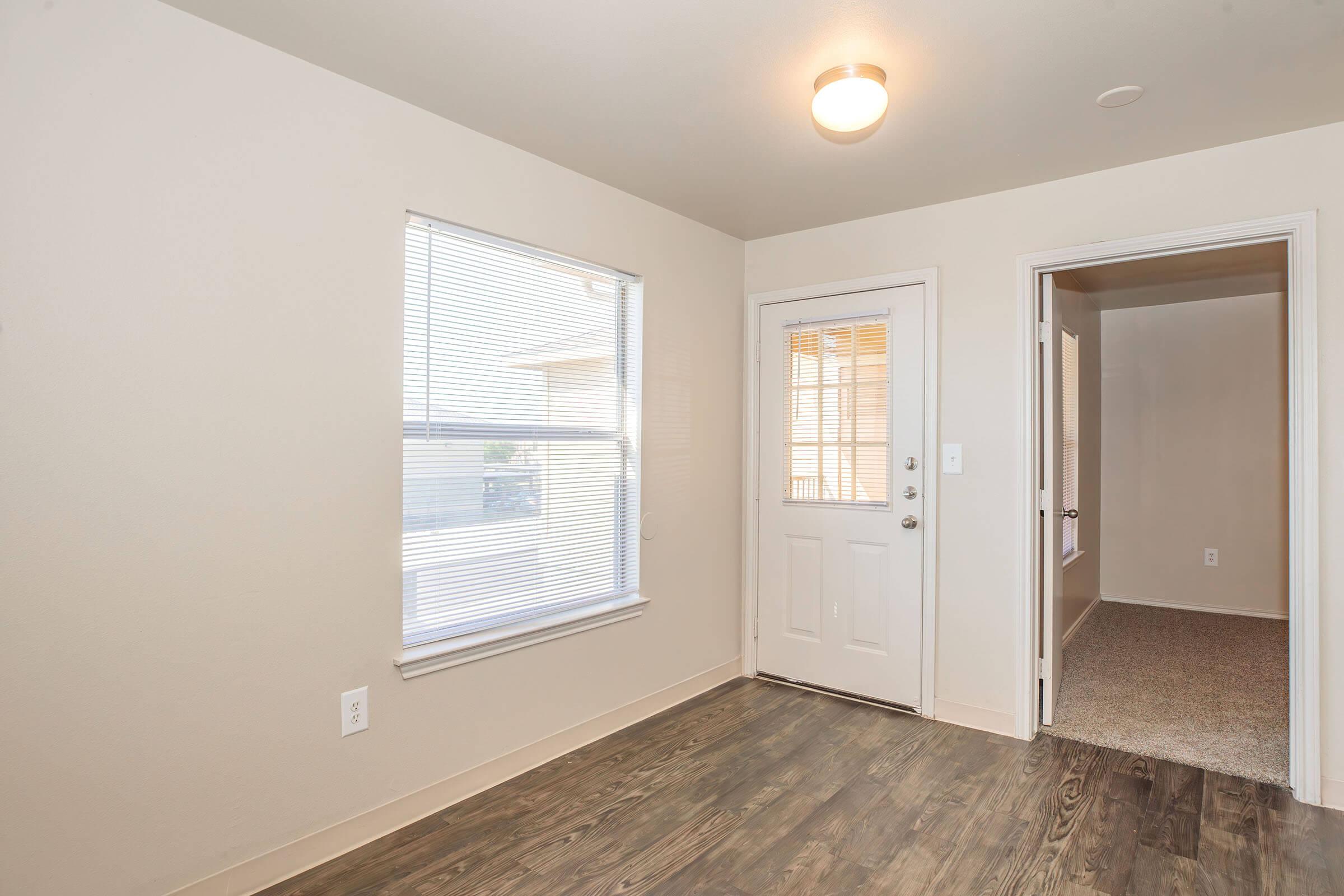 A well-lit room featuring a window with blinds on one wall and a door leading outside. The floor is a dark wood laminate, and there's a doorway leading to another room. The walls are painted a light color, creating a clean and spacious atmosphere.