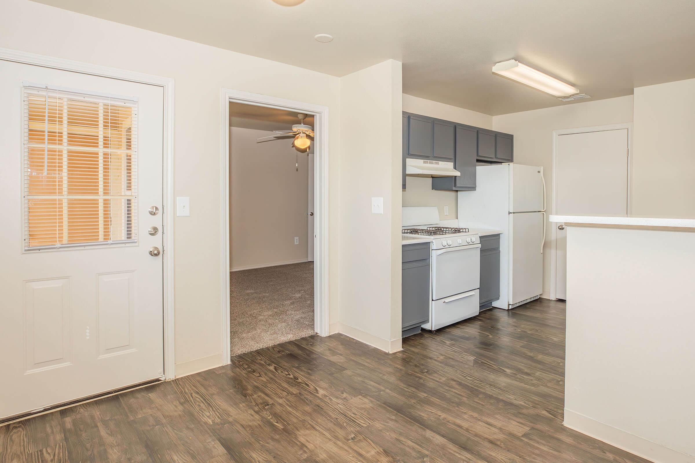Interior view of a modern apartment featuring a kitchen with gray cabinets and white appliances, a door leading outside, and a hallway leading to a carpeted living area with a ceiling fan. Light-colored walls and hardwood floors create a bright and inviting space.