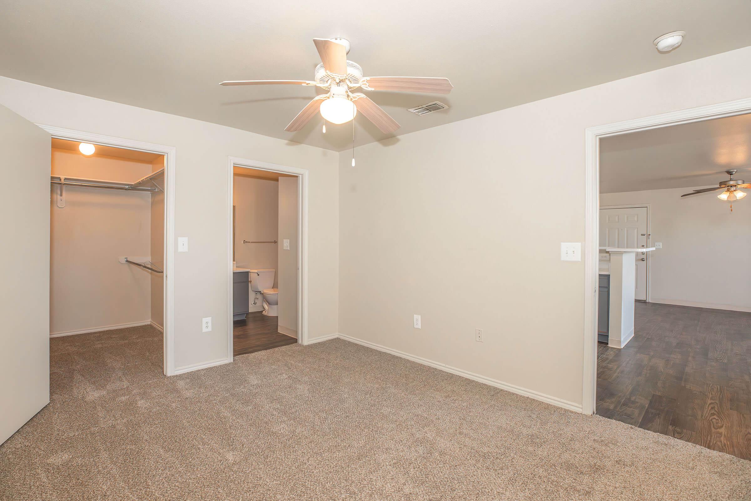 A spacious, well-lit room featuring neutral walls and carpet flooring. It includes a ceiling fan with light, doorways leading to adjoining rooms, and a closet space. The room has a clean and modern appearance, suitable for various uses, such as a bedroom or office.