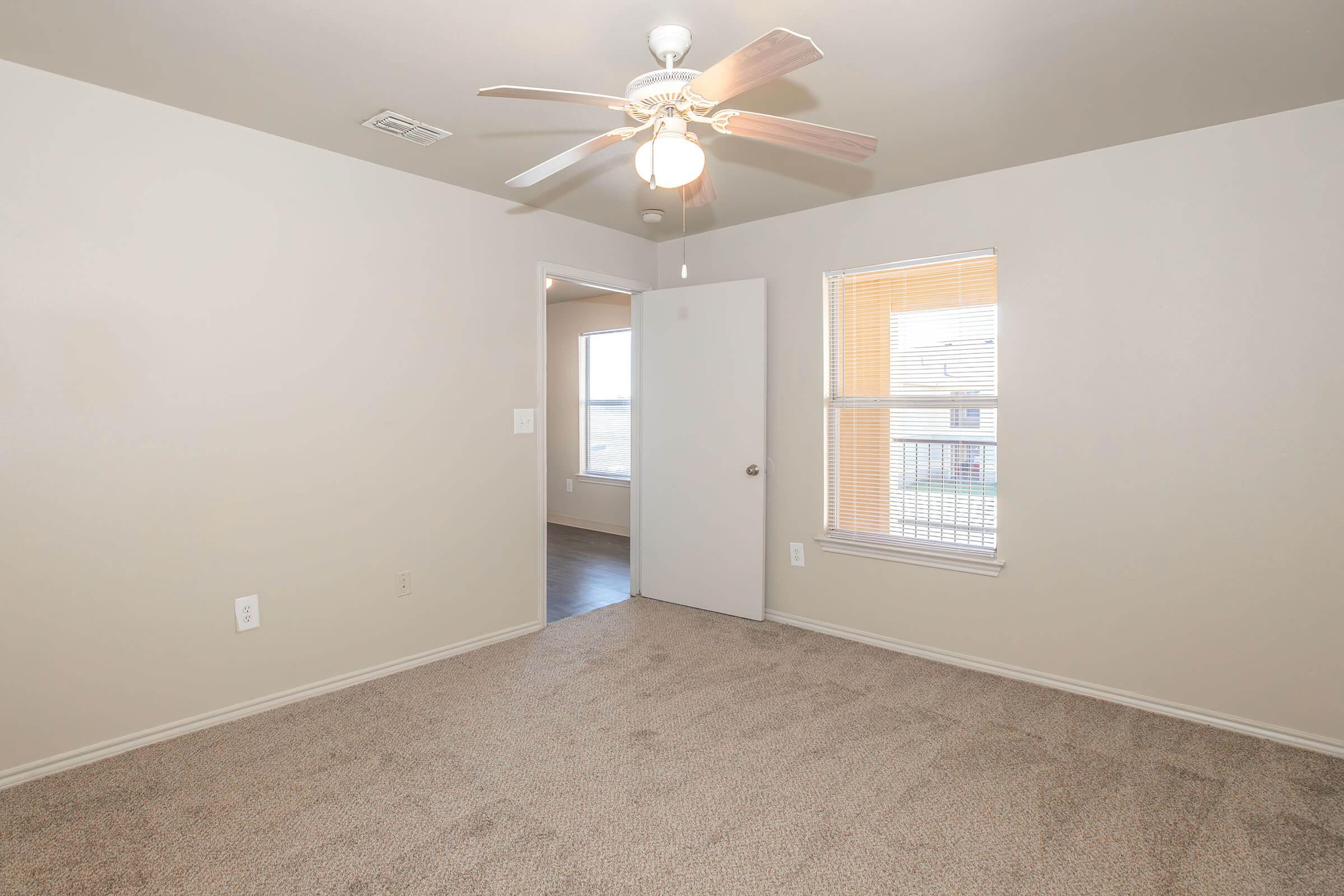 A well-lit, empty room featuring beige walls and carpeted flooring. It has a ceiling fan, an open door leading to another room, and a window allowing natural light. The overall atmosphere is tidy and spacious, suitable for personalizing and decorating.
