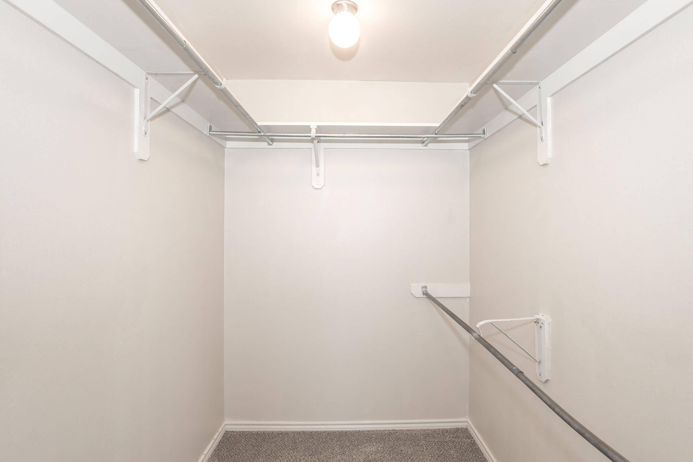 A spacious, empty closet featuring white walls, a single overhead light fixture, and two clothing rods. The floor is covered with a textured surface, and there are no visible shelves or items in the closet, creating a clean and minimalist appearance.