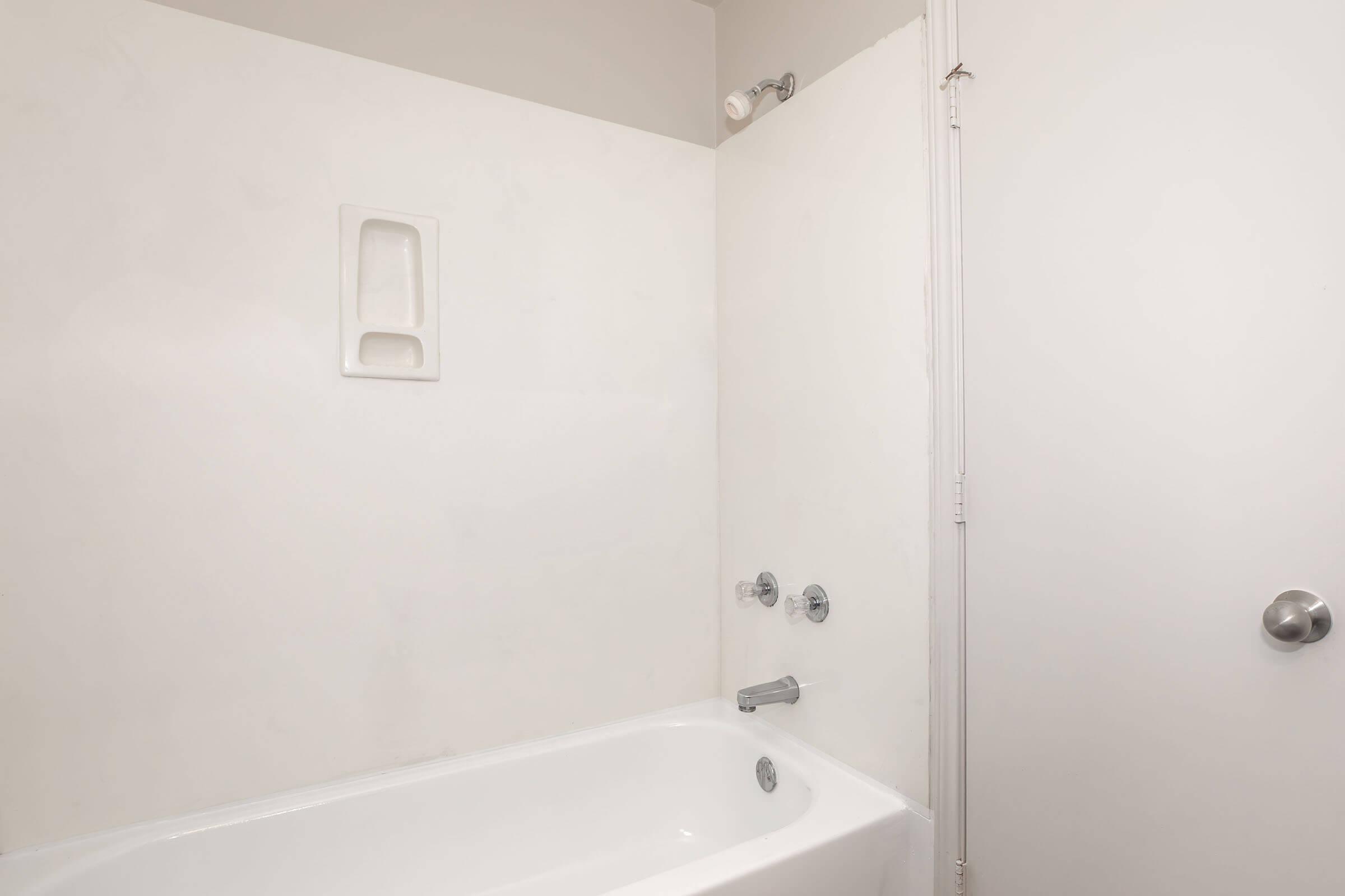A clean, minimalist bathroom scene featuring a white bathtub against a light gray wall. There is a showerhead mounted on the wall above the tub, and a small wall shelf is built into the wall for storage. A plain white door is visible, providing access to the bathroom. The overall aesthetic is simple and modern.