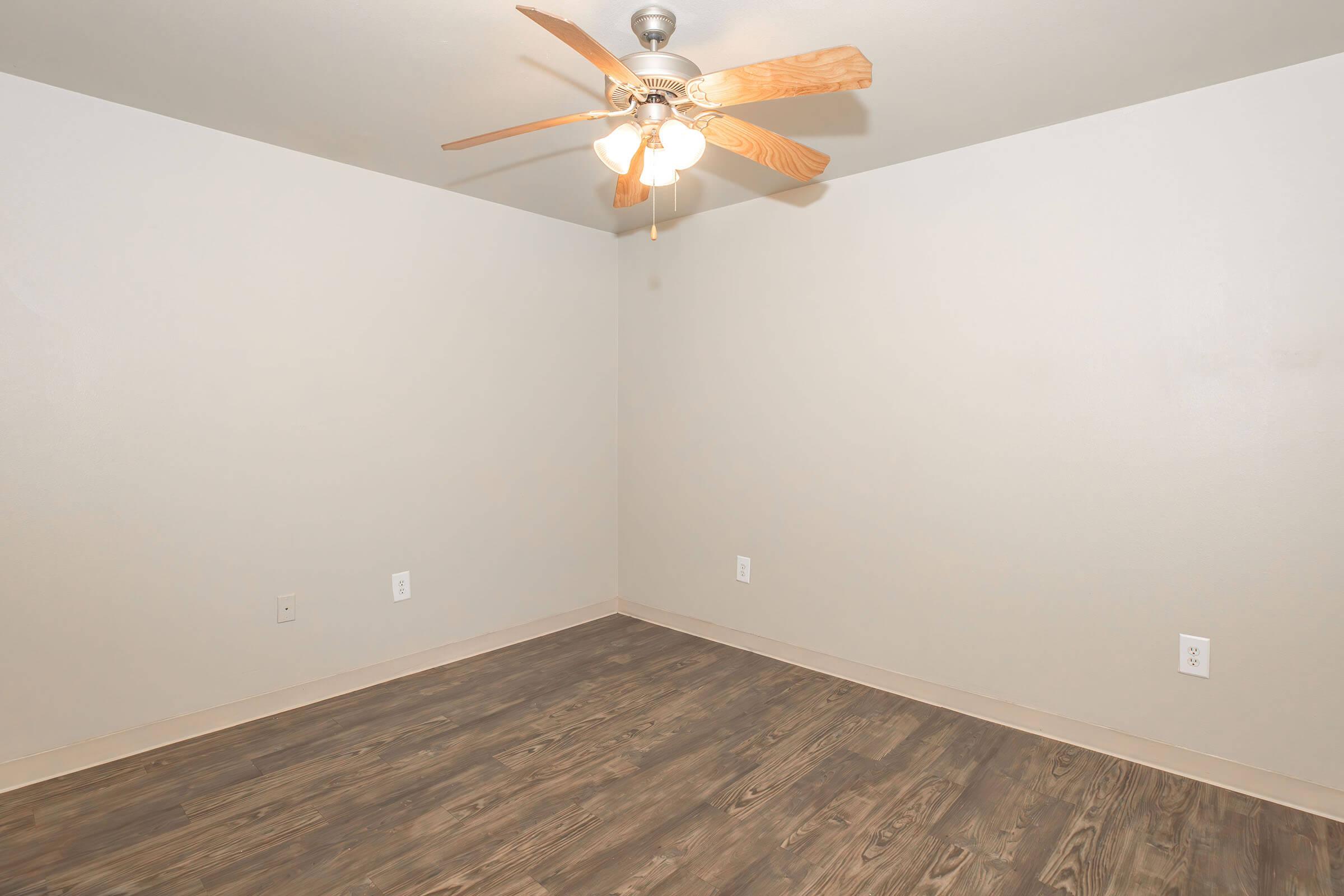 Empty room with light-colored walls and wooden flooring. A ceiling fan with wooden blades hangs from the ceiling. The space is unfurnished, creating a clean and spacious feel, ready for decoration or furniture placement.