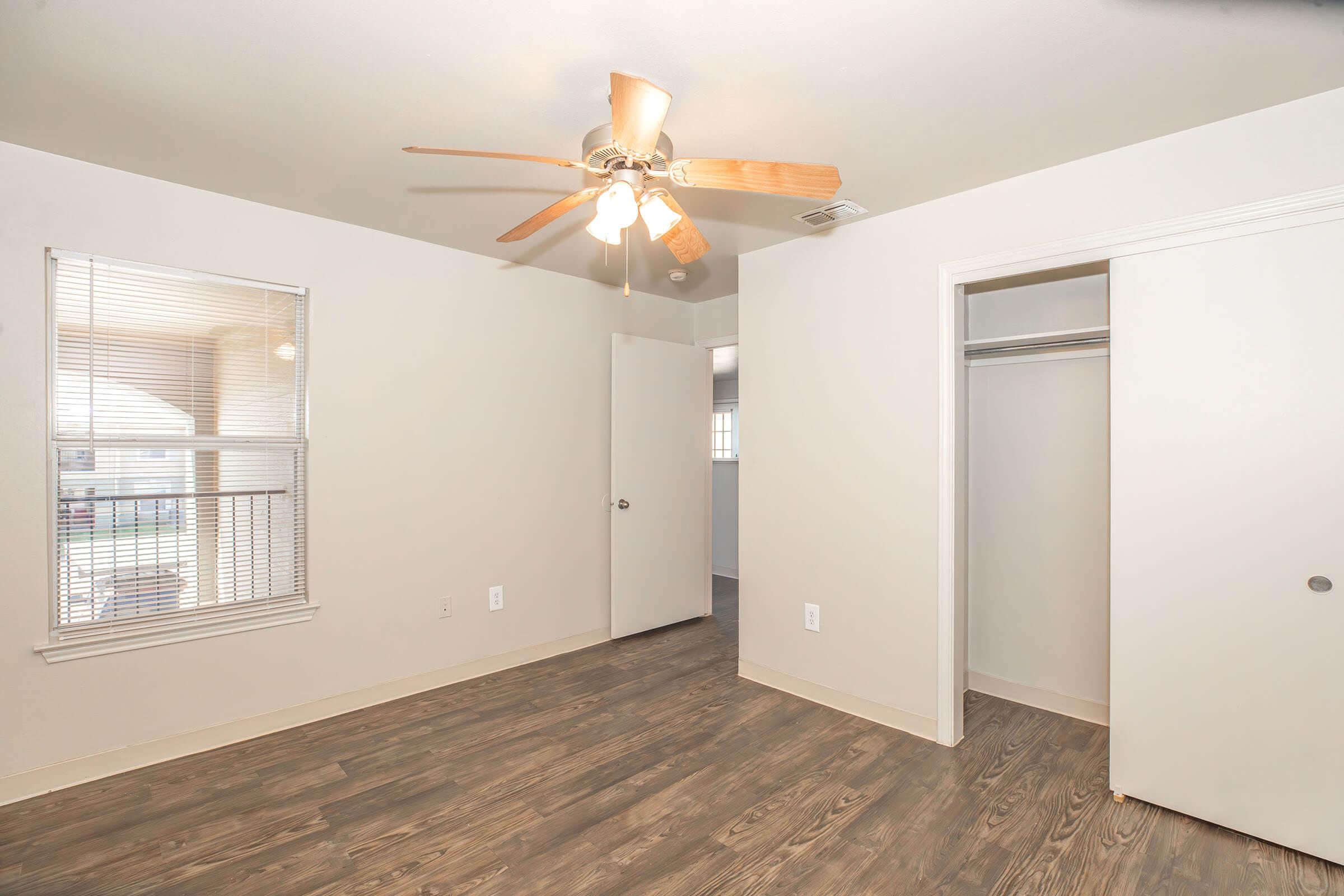 A spacious room with light-colored walls and a ceiling fan. Features include a window with blinds letting in natural light, a doorway leading to another space, and a closet with sliding doors. The floor is a warm wood-like finish, creating a cozy atmosphere.