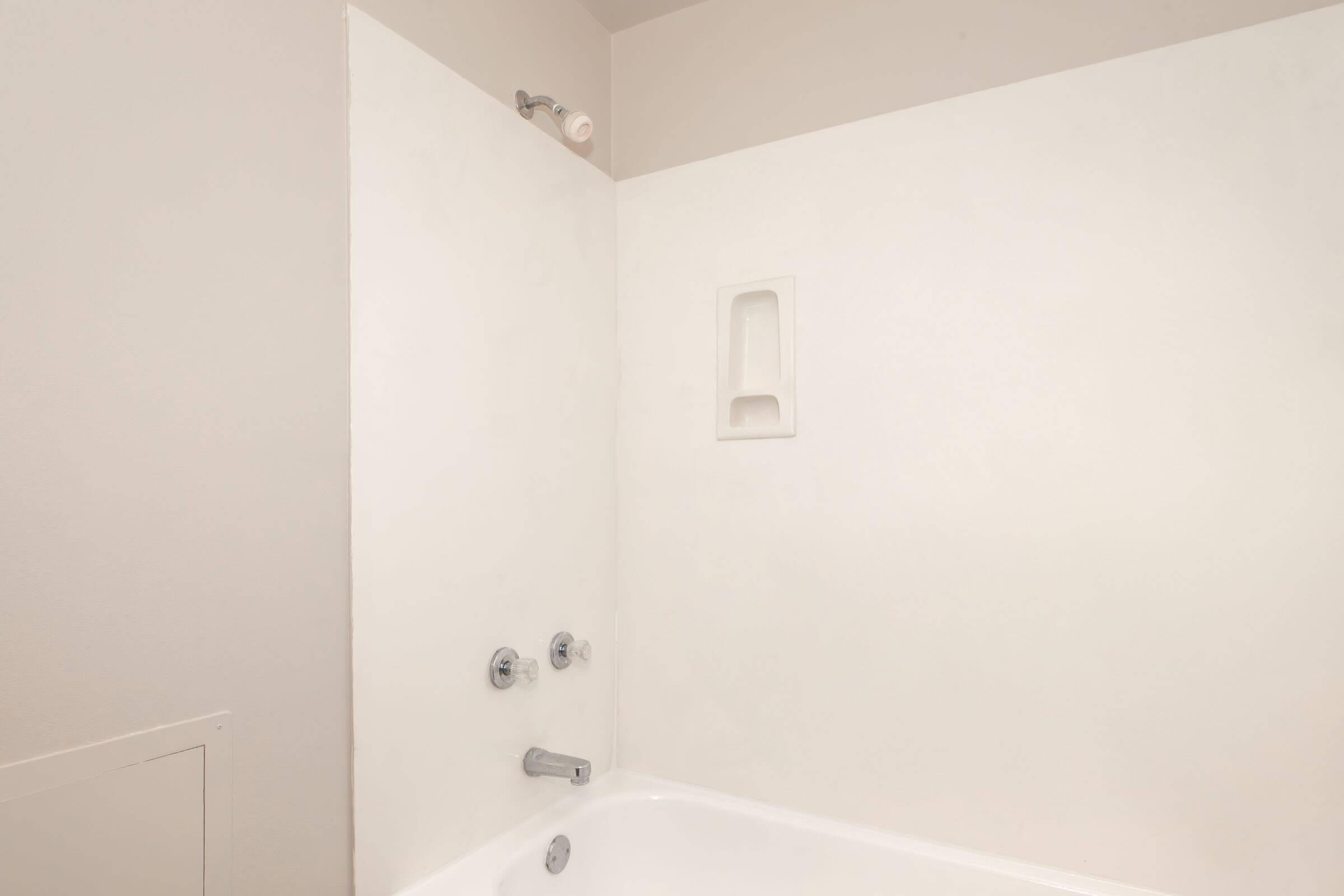 A clean, minimalist bathroom featuring a white tub and plain white walls. The showerhead is mounted on the wall, and there is a built-in shelf in the corner. The space is bright and uncluttered, emphasizing a simple design.