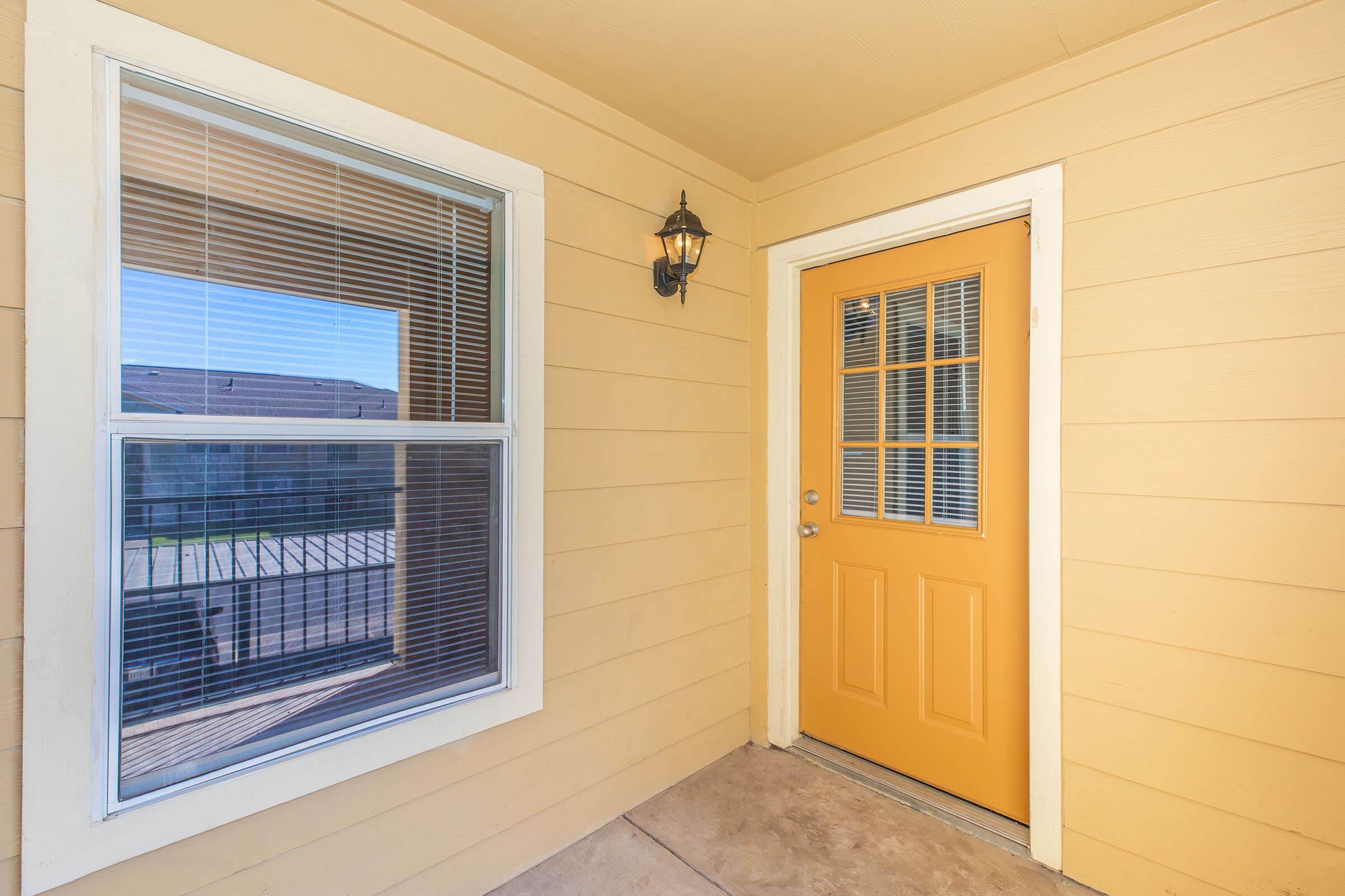 A well-lit entryway featuring a bright yellow door with multiple panes, situated between two windows. The walls are painted a light yellow, and there is a wall-mounted light fixture beside the door, adding to the welcoming appearance. The floor is made of concrete, leading to an outdoor area.