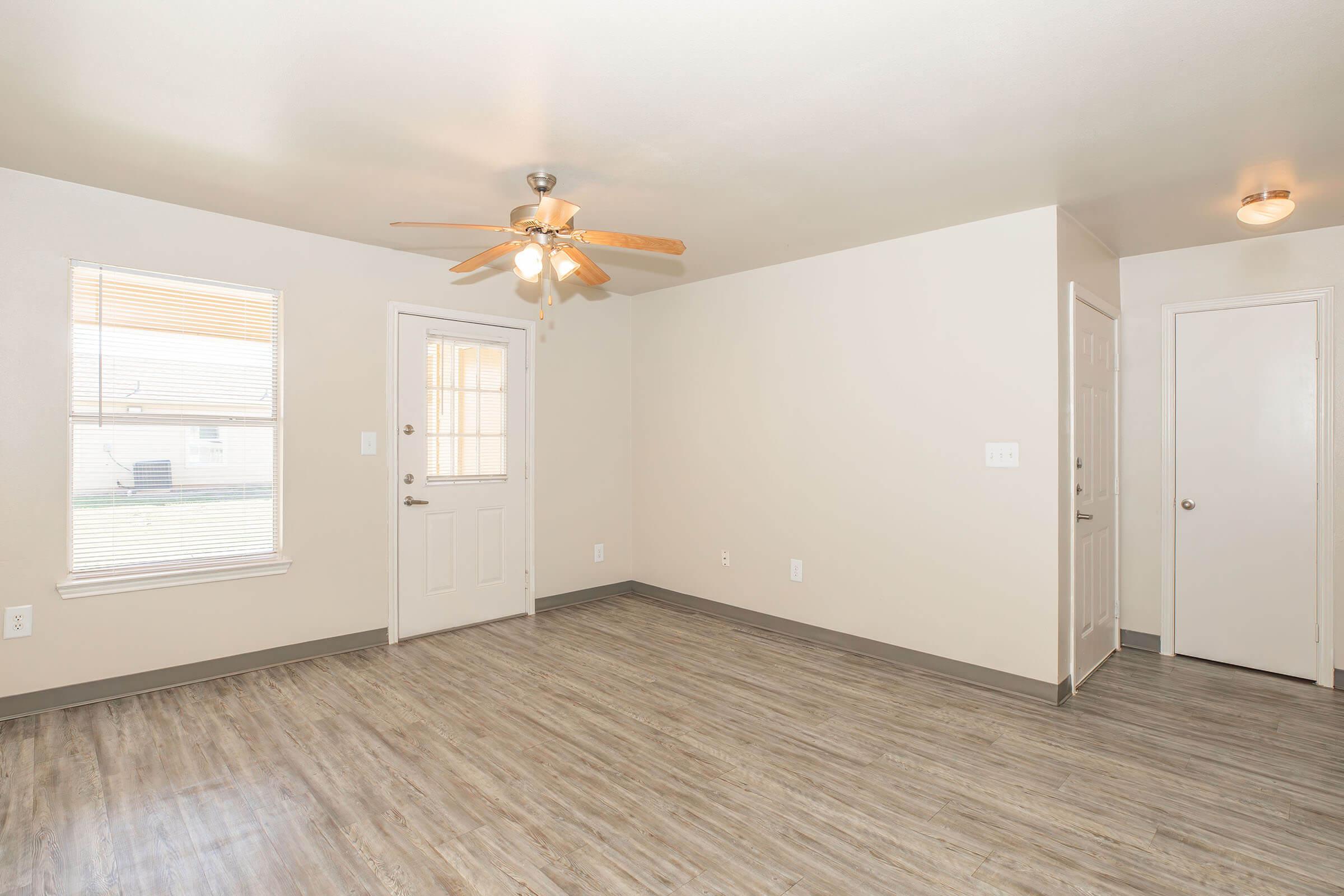 An empty room featuring a ceiling fan and light fixture, a front door with a window, and an adjacent door leading to another area. The floor is covered in light-colored laminate, and there are two windows allowing natural light to enter the space. The walls are painted in a neutral tone.