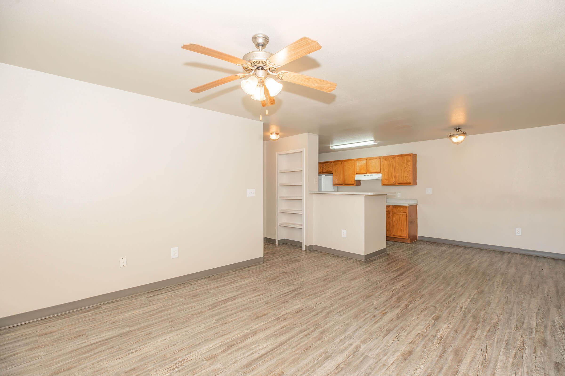 Spacious interior of an apartment featuring a ceiling fan, light-colored walls, and wooden cabinets in the kitchen area. The flooring is light wood-like laminate, and there are built-in shelves visible near the kitchen. The room is well-lit and offers an open layout.