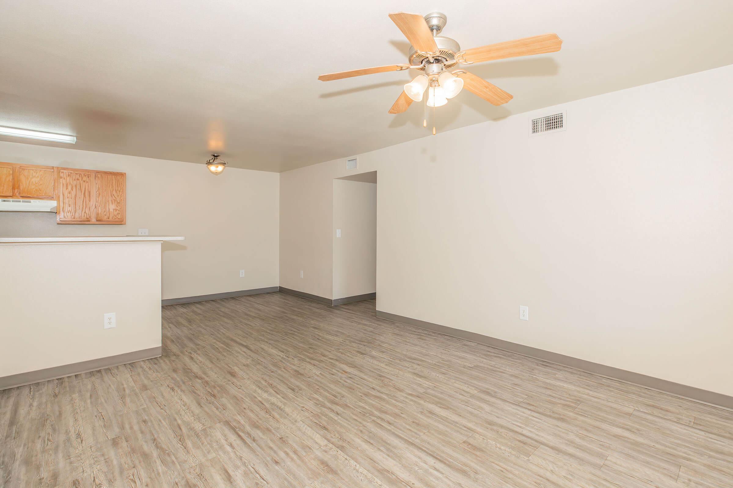 A spacious living area featuring light-colored walls, a ceiling fan with wooden blades, and a laminate floor. The kitchen is partially visible with wooden cabinetry in the background. The room has a neutral palette and large open space, ideal for furniture arrangement.