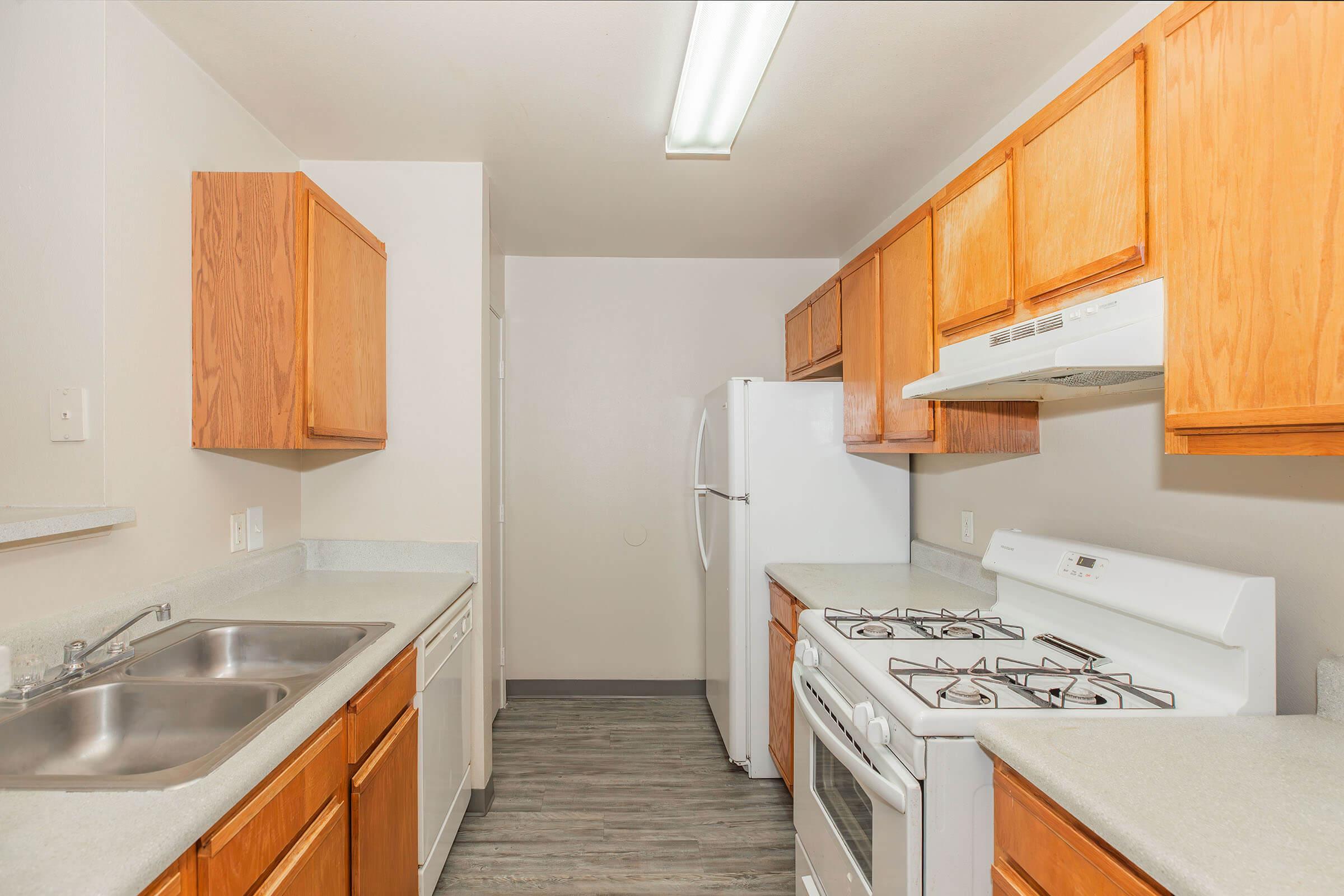 A modern kitchen featuring light wood cabinets, a double sink, a white refrigerator, and a gas stove. The countertops are light-colored, and the flooring is a sleek gray. The space is well-lit with overhead lighting, providing a clean and inviting atmosphere.