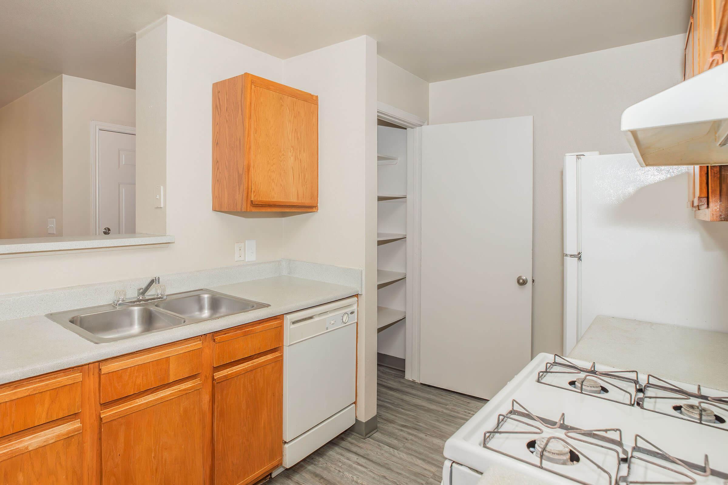 A clean, modern kitchen featuring wooden cabinets, a double sink, a dishwasher, a gas stove, and a refrigerator. There is a pantry door and light-colored walls, with a gray floor providing a contemporary look. The kitchen is well-lit and spacious, emphasizing functionality and comfort.