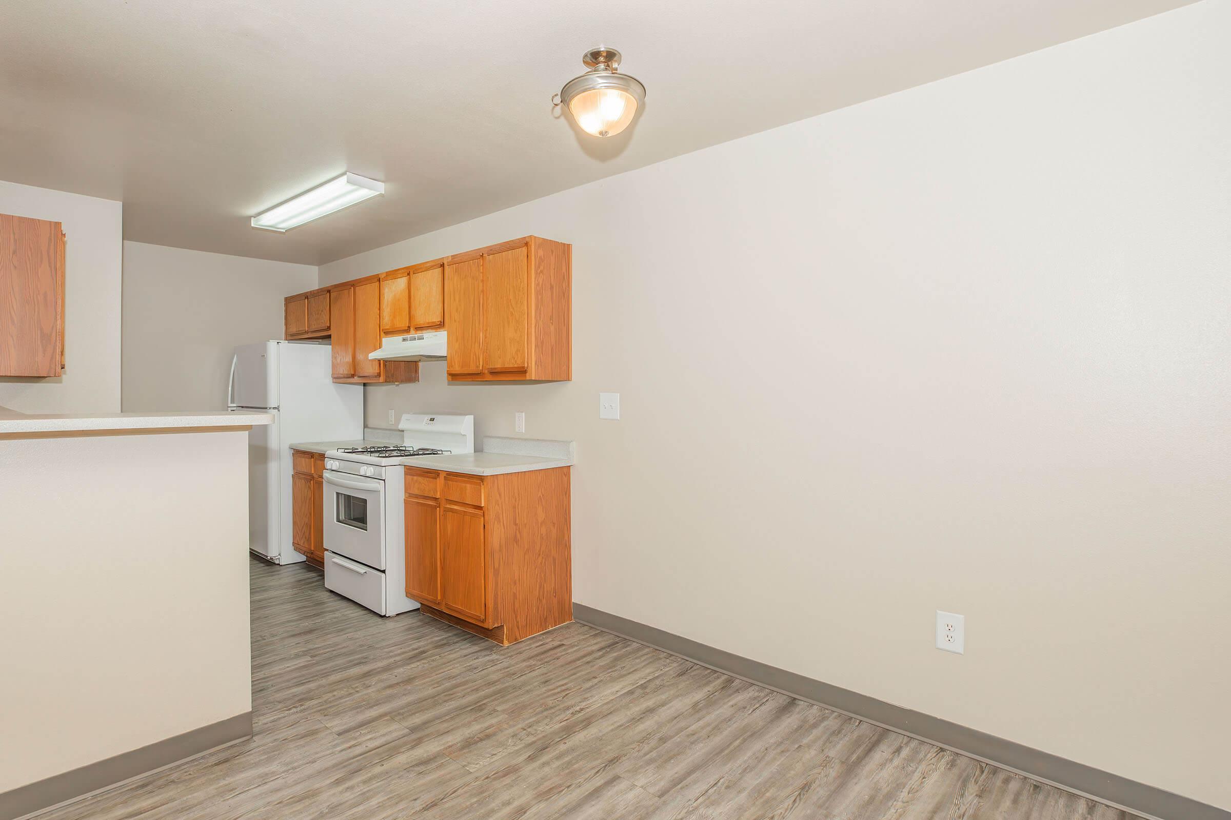 A modern kitchen in an apartment, featuring wooden cabinetry, a white gas stove, and a refrigerator. The floor has a wood-like finish, and there is a ceiling light fixture. The walls are painted in light color, creating a bright and spacious feel.