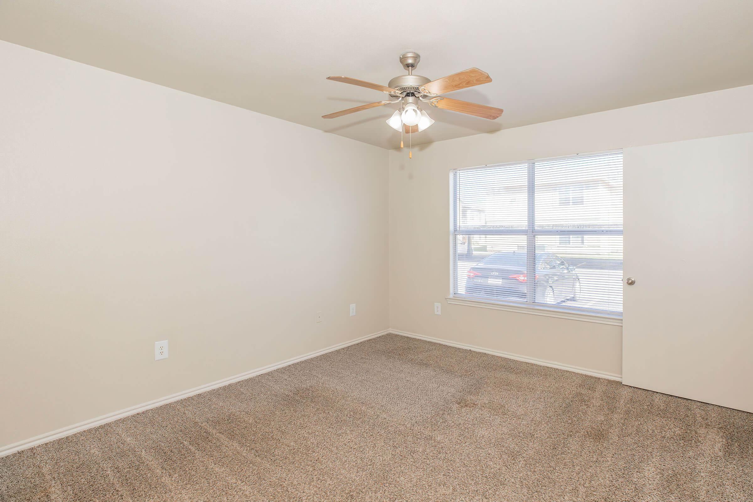A spacious, empty room with beige walls and light brown carpet. A ceiling fan with wooden blades is mounted in the center. A large window lets in natural light, showing a glimpse of a parked car outside. The room features a door on the right side leading to another area.