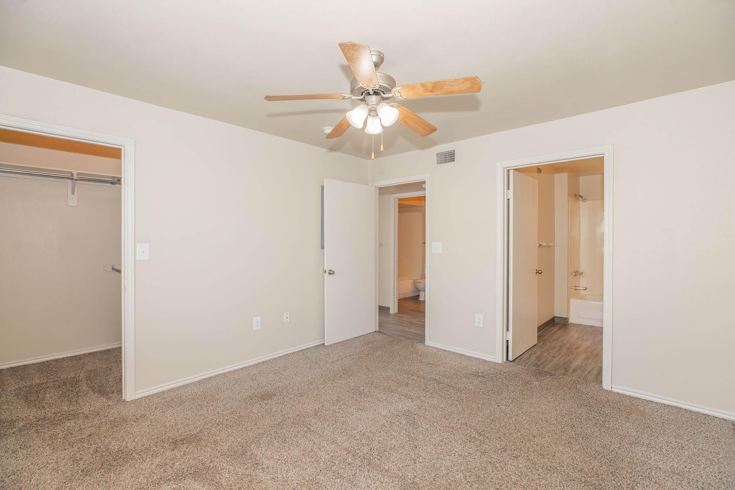 A spacious room with light-colored walls and carpet flooring. A ceiling fan with wooden blades is installed. There are two doorways leading to adjacent rooms, one with a built-in closet and another leading to a bathroom. Natural light illuminates the room, creating a warm and inviting atmosphere.