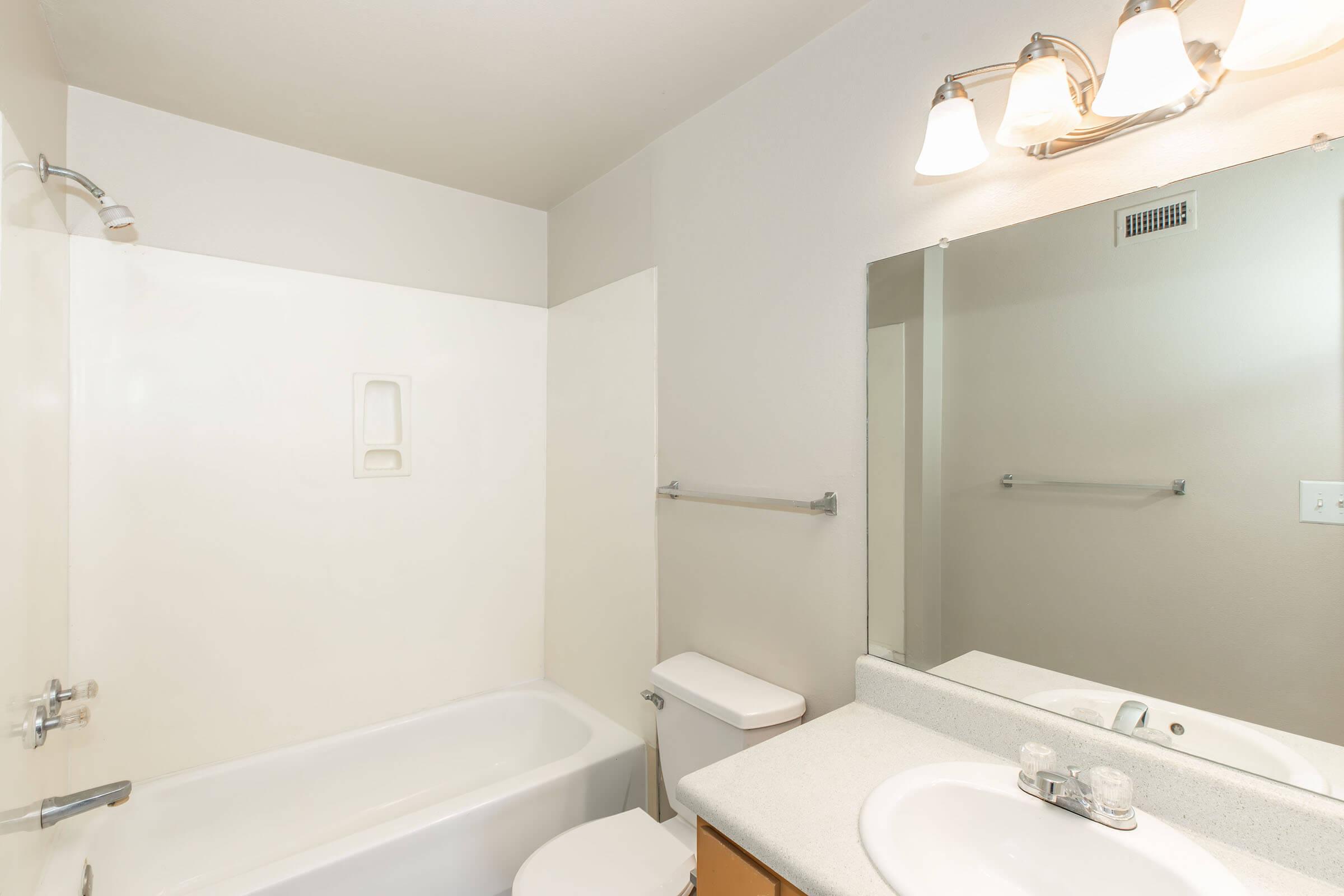 A clean and minimalistic bathroom featuring a white bathtub, a toilet, and a sink with a countertop. The walls are mostly white with a light-colored accent. Above the sink, there is a mirror and a light fixture. A towel rack is mounted on the side wall. The space is well-lit and organized.