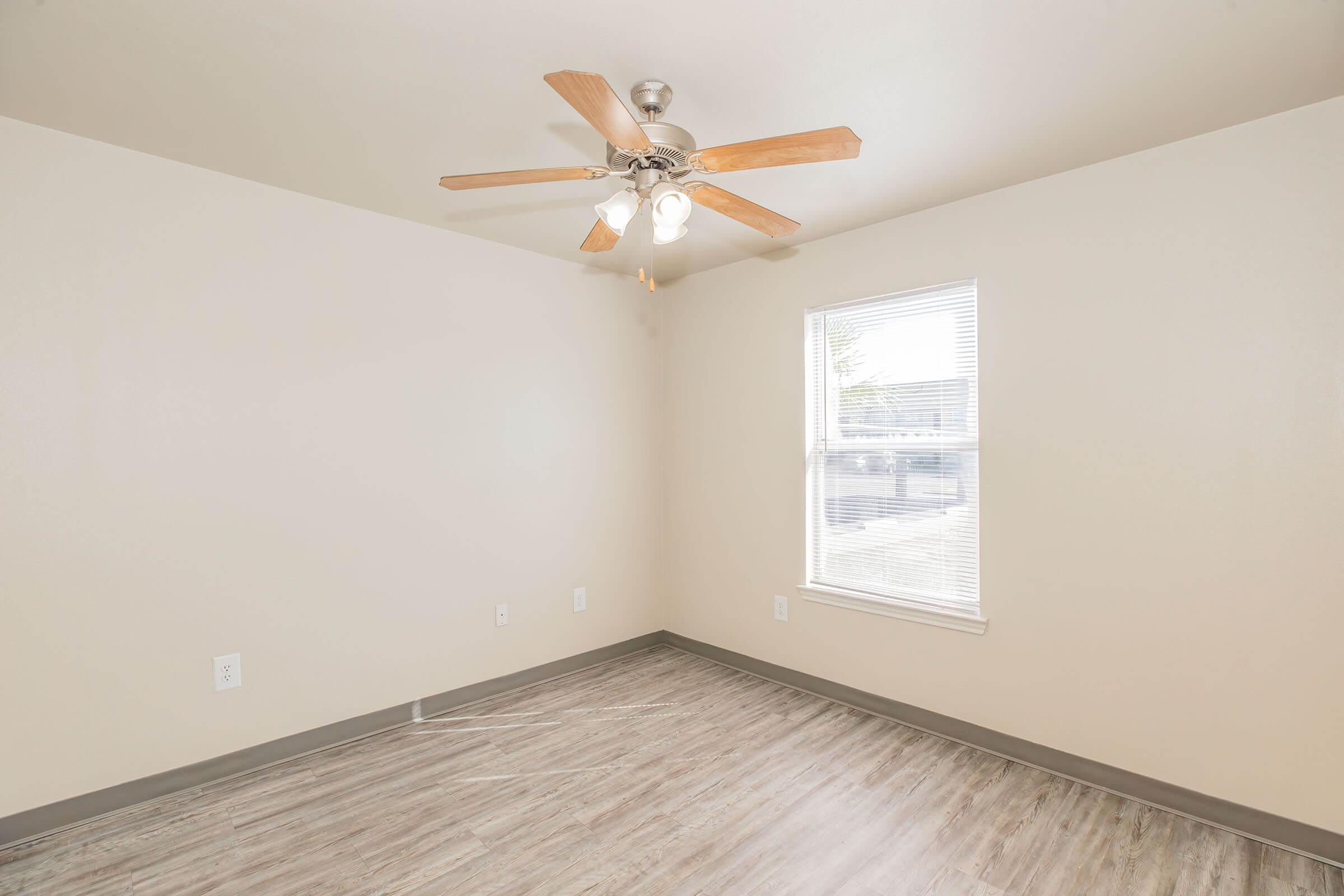 A well-lit, empty room with light-colored walls and a ceiling fan with wooden blades. The floor features light-colored laminate, and there is a single window allowing natural light to enter. The room has a clean, minimalist appearance, suitable for various uses.