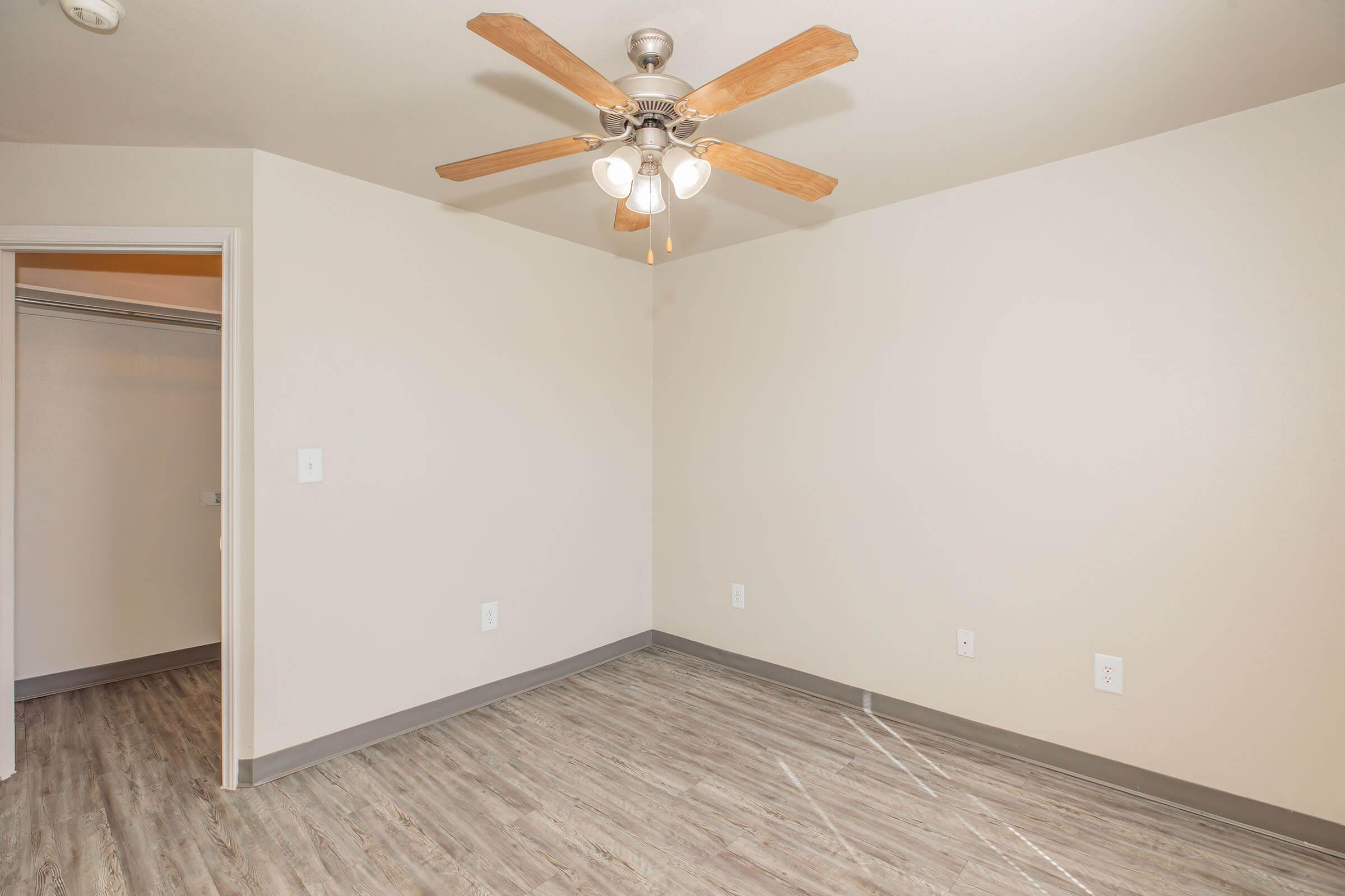 A well-lit empty room featuring a ceiling fan with wooden blades. The walls are painted a light color, and the flooring is a light wood laminate. A closet is visible on the left side of the image, showcasing its spaciousness and minimalistic design, creating a versatile space for various uses.