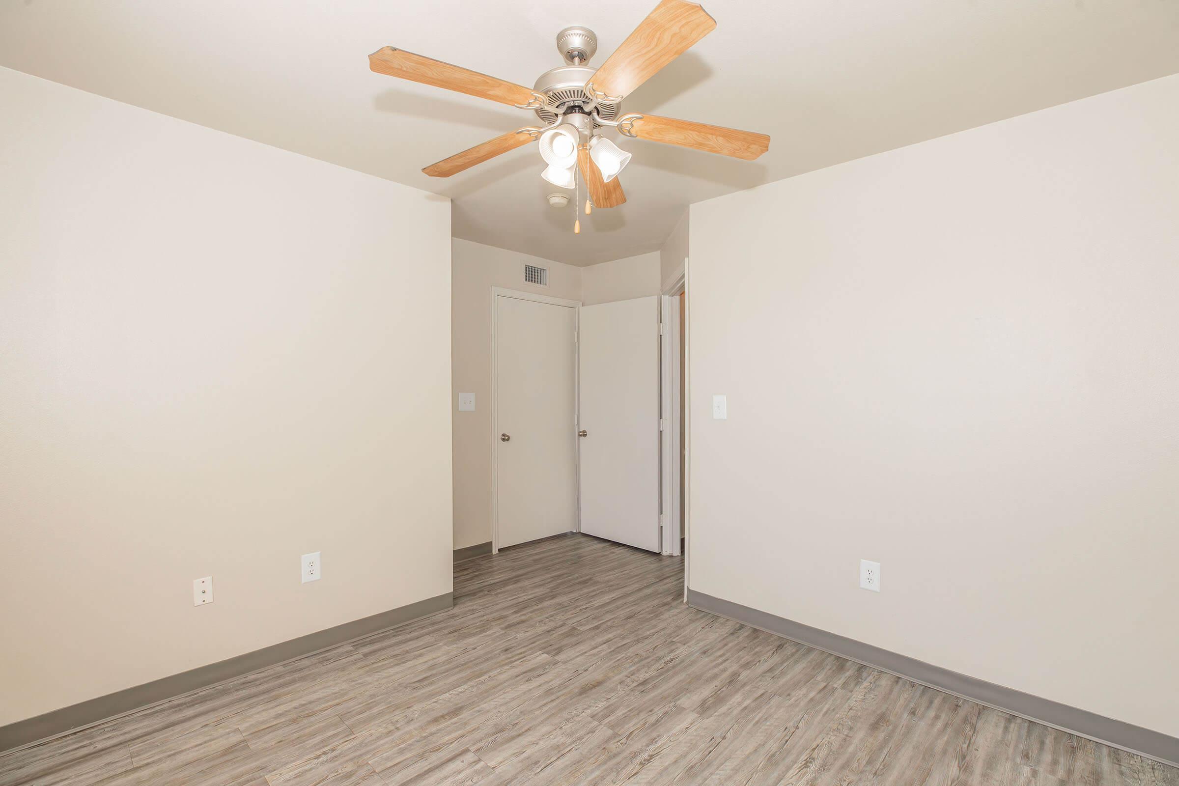 An empty room featuring light gray walls and a ceiling fan with wooden blades. The floor is a light wood-like laminate. A closed door is visible on the left, and another door is partially open on the right, suggesting access to another space.