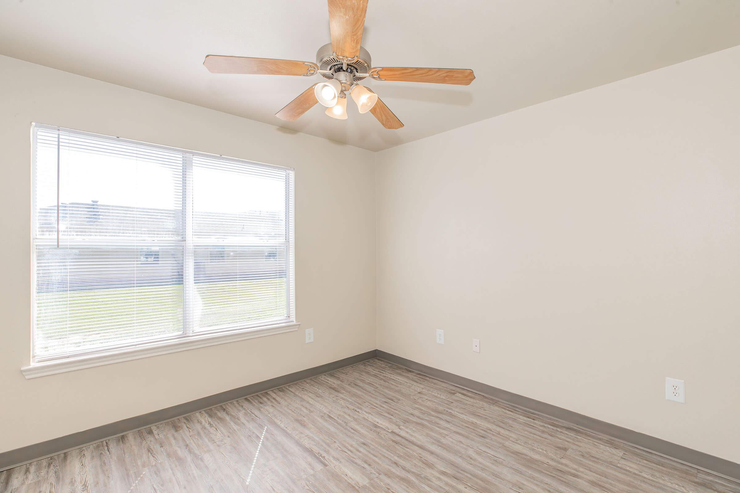 Empty room with light-colored walls, a ceiling fan with wooden blades, and a large window allowing natural light to enter. The floor features light wood-like laminate, and there is a grassy area visible outside the window. The overall space appears bright and airy.