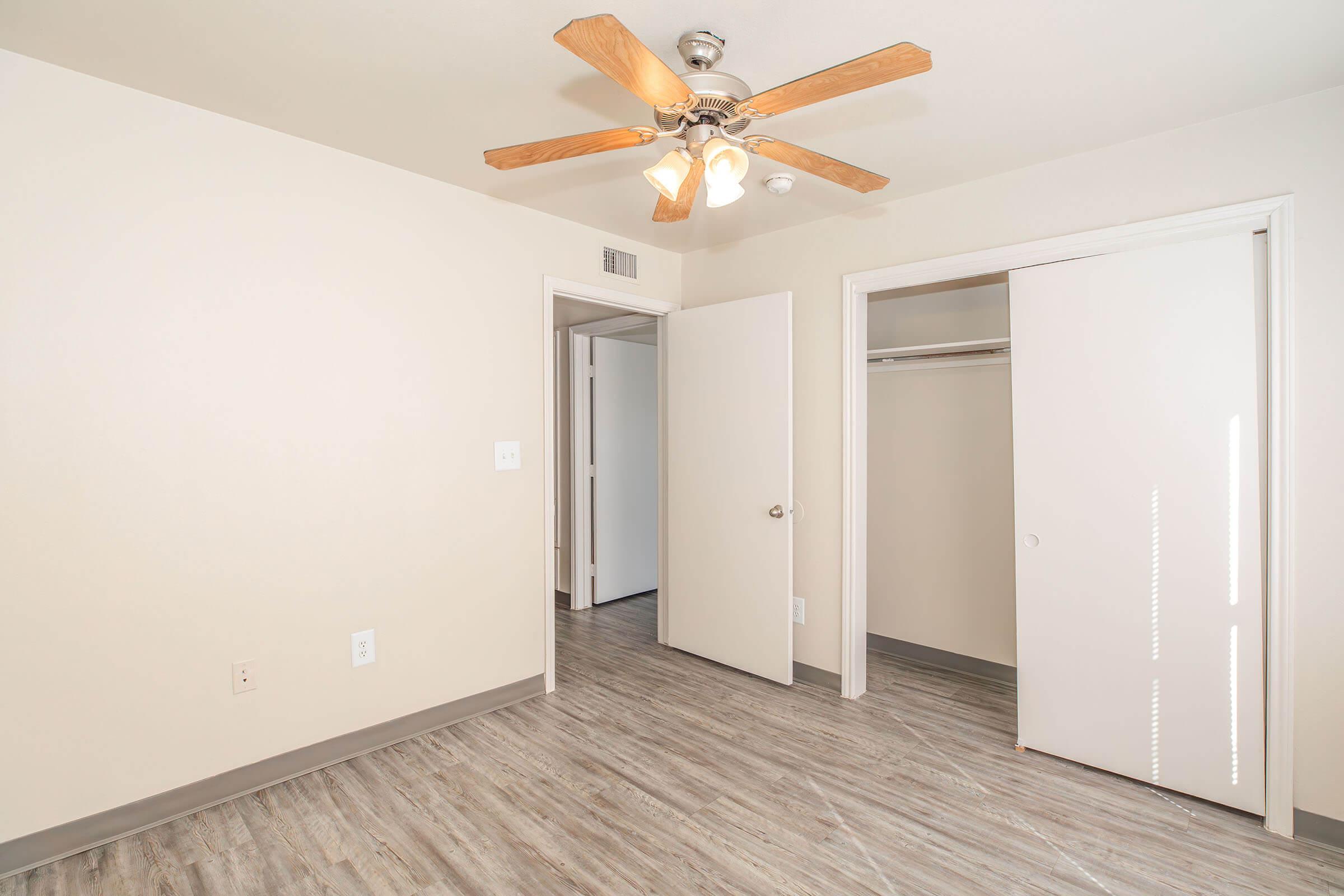 A light-colored room featuring a ceiling fan with wooden blades, a closet with sliding doors, and a light-colored wall. The floor is covered with plank-style flooring, and there are two doorways leading to other areas. The overall atmosphere is bright and spacious.