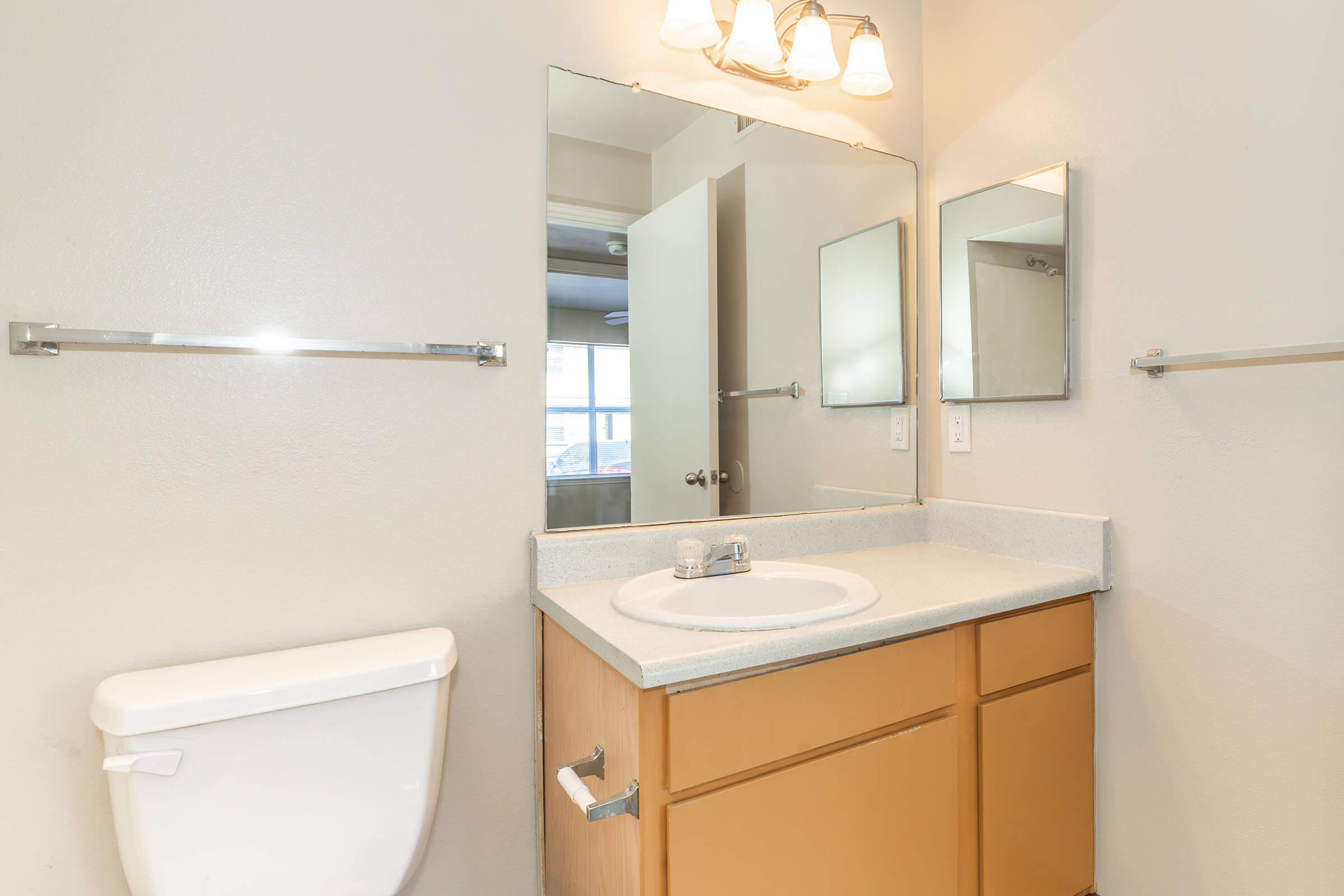A clean bathroom featuring a single sink with a countertop, a large mirror above, and a light fixture with three bulbs. To the left, there's a toilet, and on the opposite wall, a partially open door leads to another room. The walls are painted in a light color, creating a bright and spacious feel.