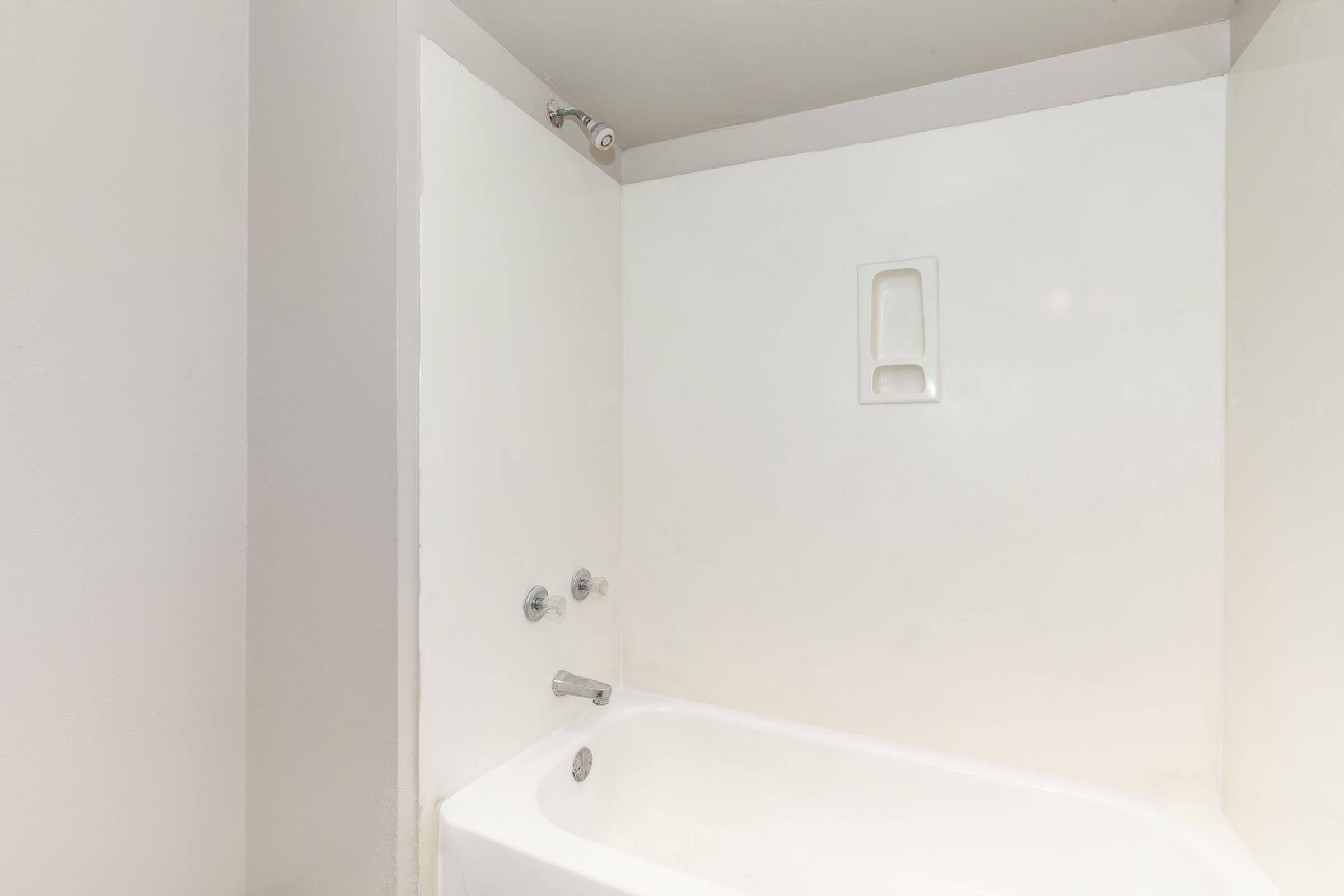 A clean, minimalist bathroom featuring a white bathtub and walls. The bathtub has a simple faucet and shower head mounted on the wall. A small niche is visible on the back wall, providing a space for toiletries. The overall design is modern and uncluttered, emphasizing functionality.