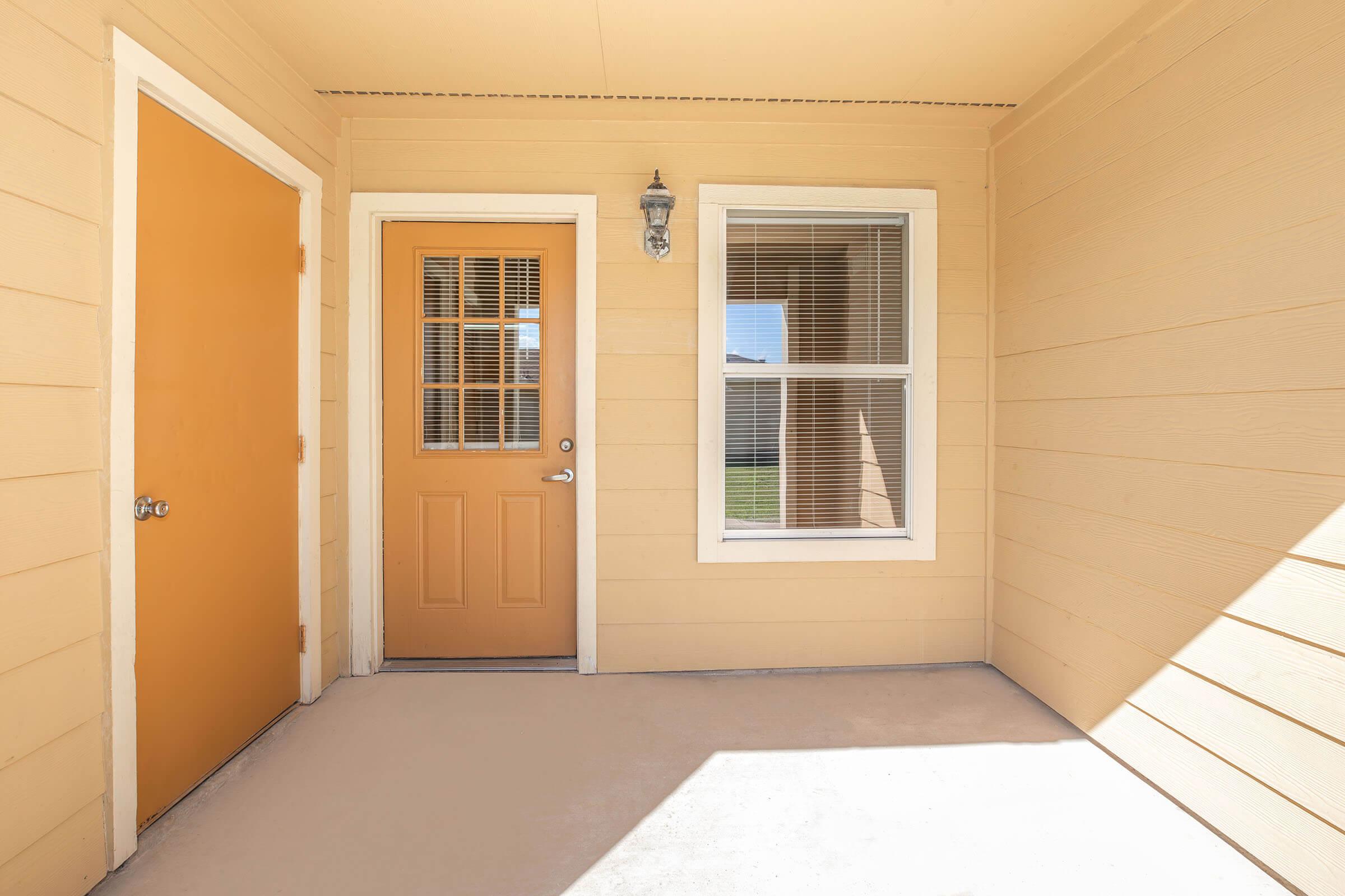 A well-lit entrance area featuring a beige exterior wall. To the left, there is a solid orange door, and to the right, a door with a window panel. A large window beside the door allows natural light to illuminate the space, which has a concrete floor. The overall atmosphere is inviting and cozy.