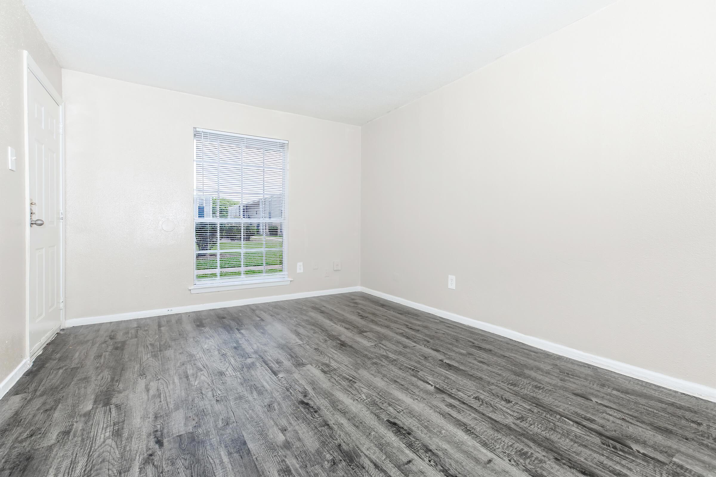 A vacant interior room with light beige walls and a medium-colored wood-like floor. A window with blinds allows natural light in, showcasing a view of greenery outside. The space is unadorned, creating a minimalist and open atmosphere.
