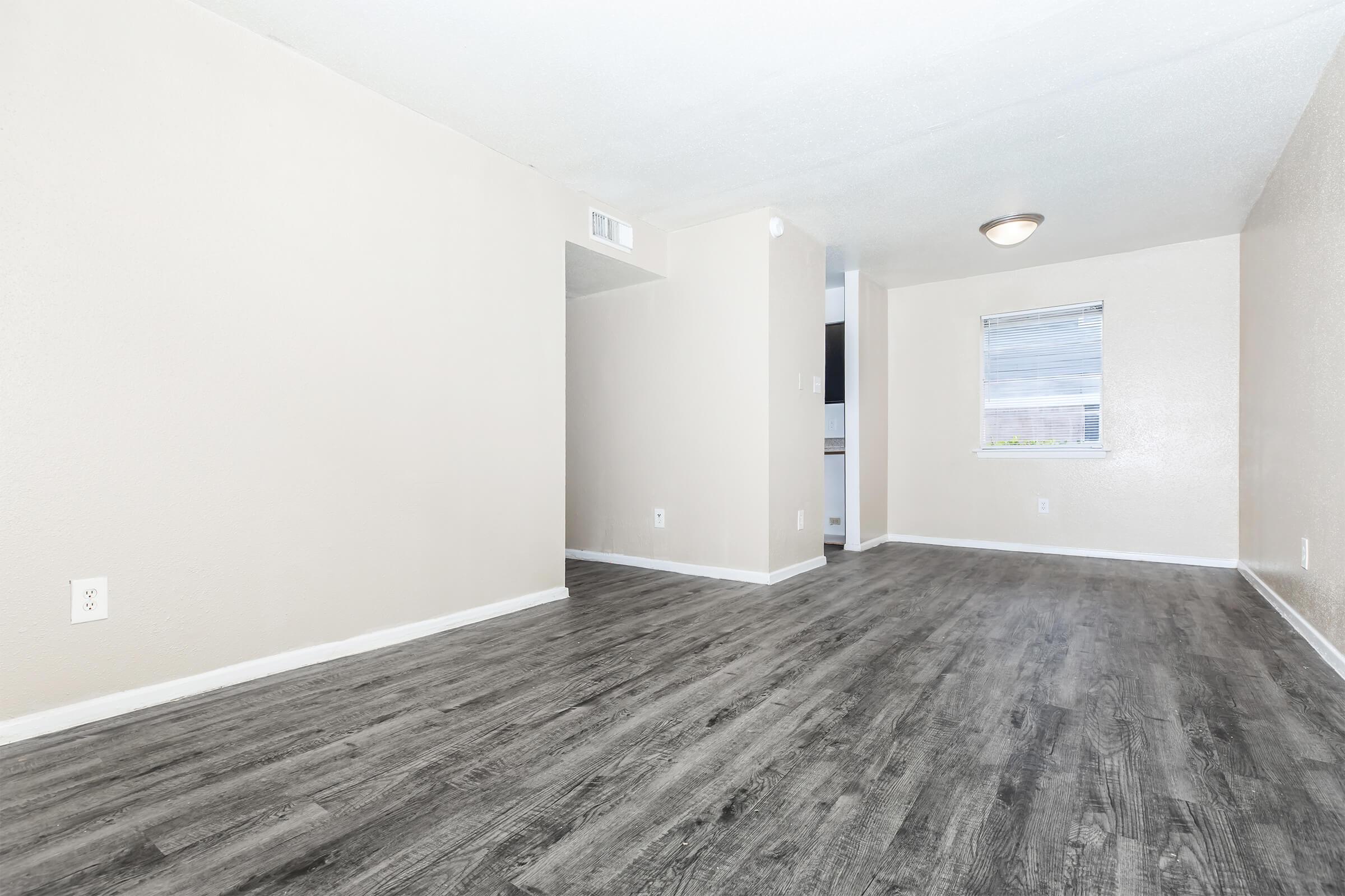 A spacious, empty living area featuring light beige walls and dark wooden laminate flooring. A single ceiling light fixture illuminates the room, which has a window providing natural light. There is a doorway leading to another space, creating an open and inviting atmosphere.