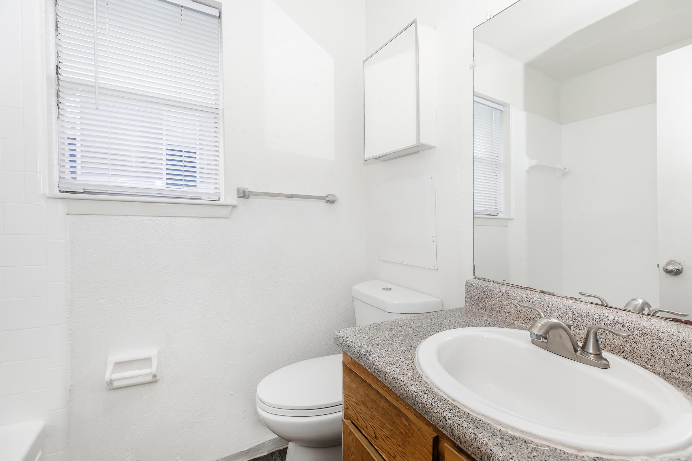 A clean, modern bathroom featuring a white wall, a mirror above a sink with a granite countertop, a toilet, and a small window with blinds. The room is well-lit and has a neutral color scheme, creating a fresh and spacious appearance.