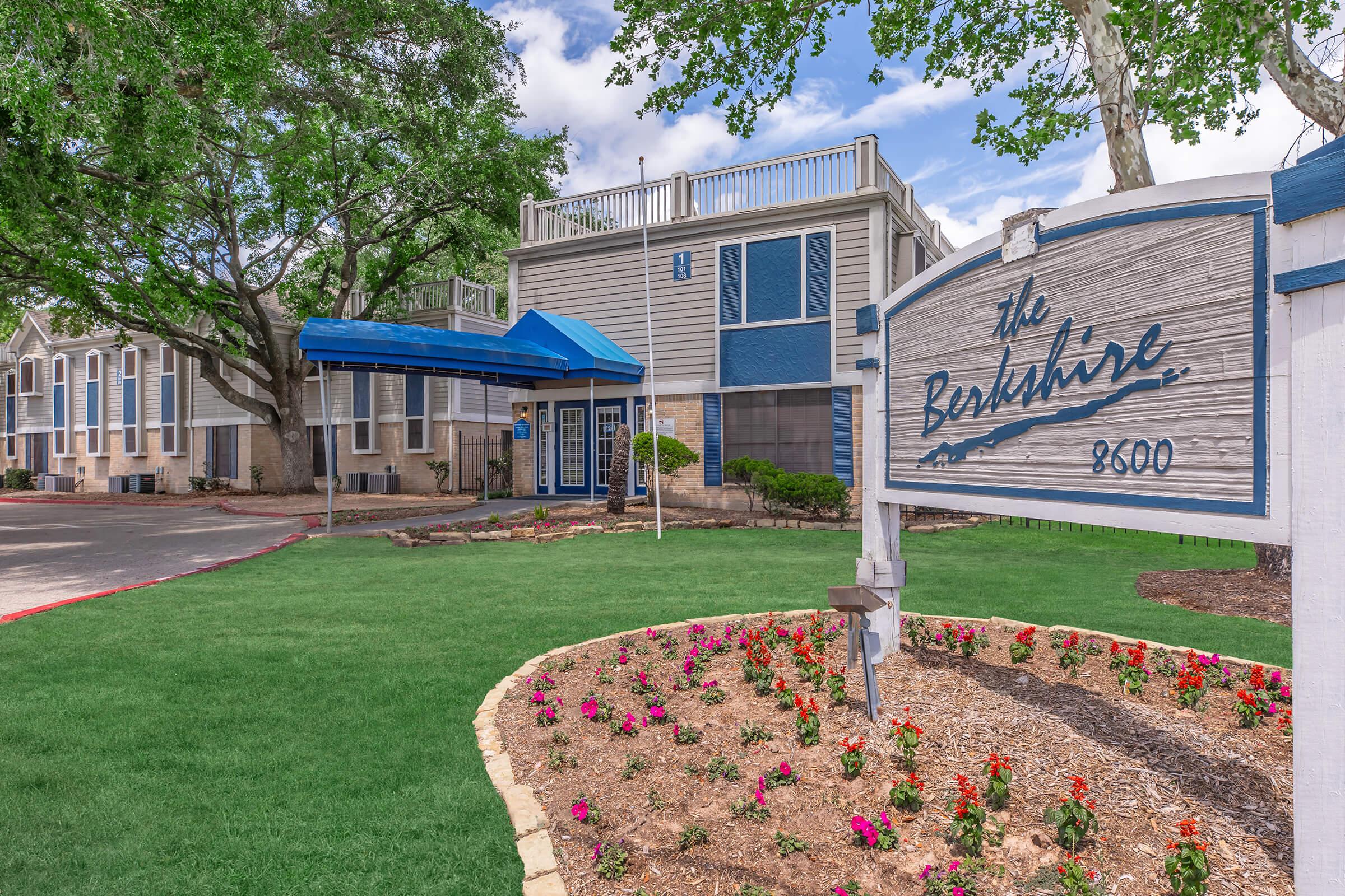 Exterior view of The Berkshire, a multi-story building featuring a blue awning and landscaped gardens with flowers. The sign prominently displays "The Berkshire" with the address 8600, set against a backdrop of trees and greenery. The sky is partly cloudy, adding to the pleasant atmosphere.