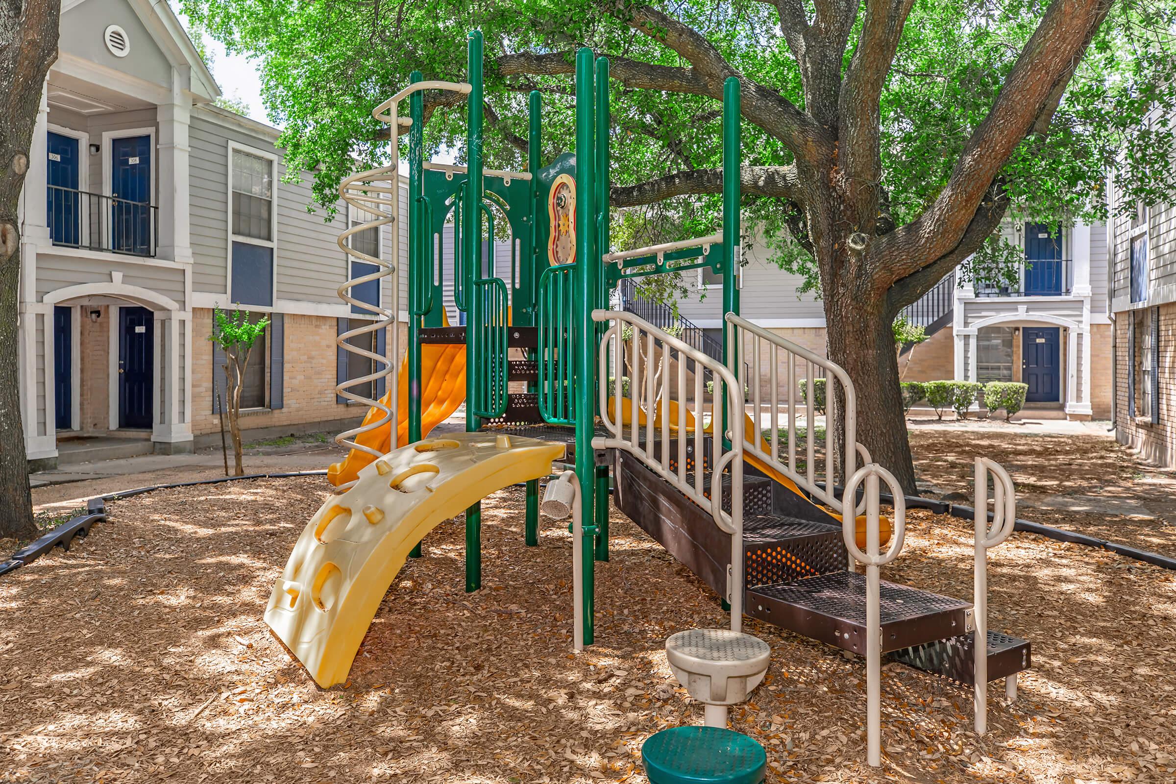 A colorful children's playground featuring a slide, climbing structures, and a spiral slide, surrounded by a soft mulch surface and shaded by large trees. The playground is set within a residential courtyard, with apartment buildings visible in the background.