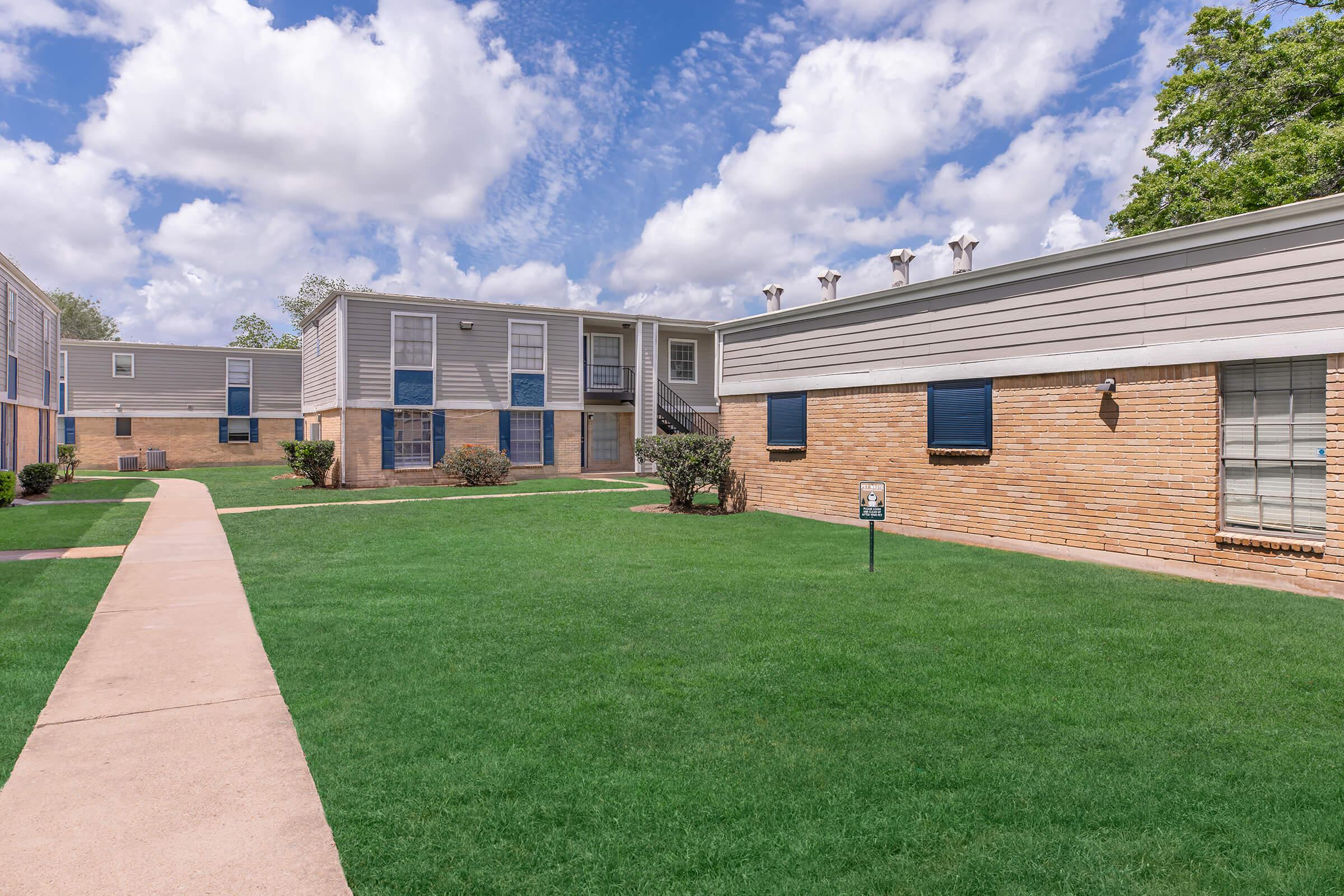 A well-maintained apartment complex featuring two buildings, surrounded by green grass and small shrubs. A paved walkway runs through the lawn, leading to the entrance of the buildings under a partly cloudy blue sky.