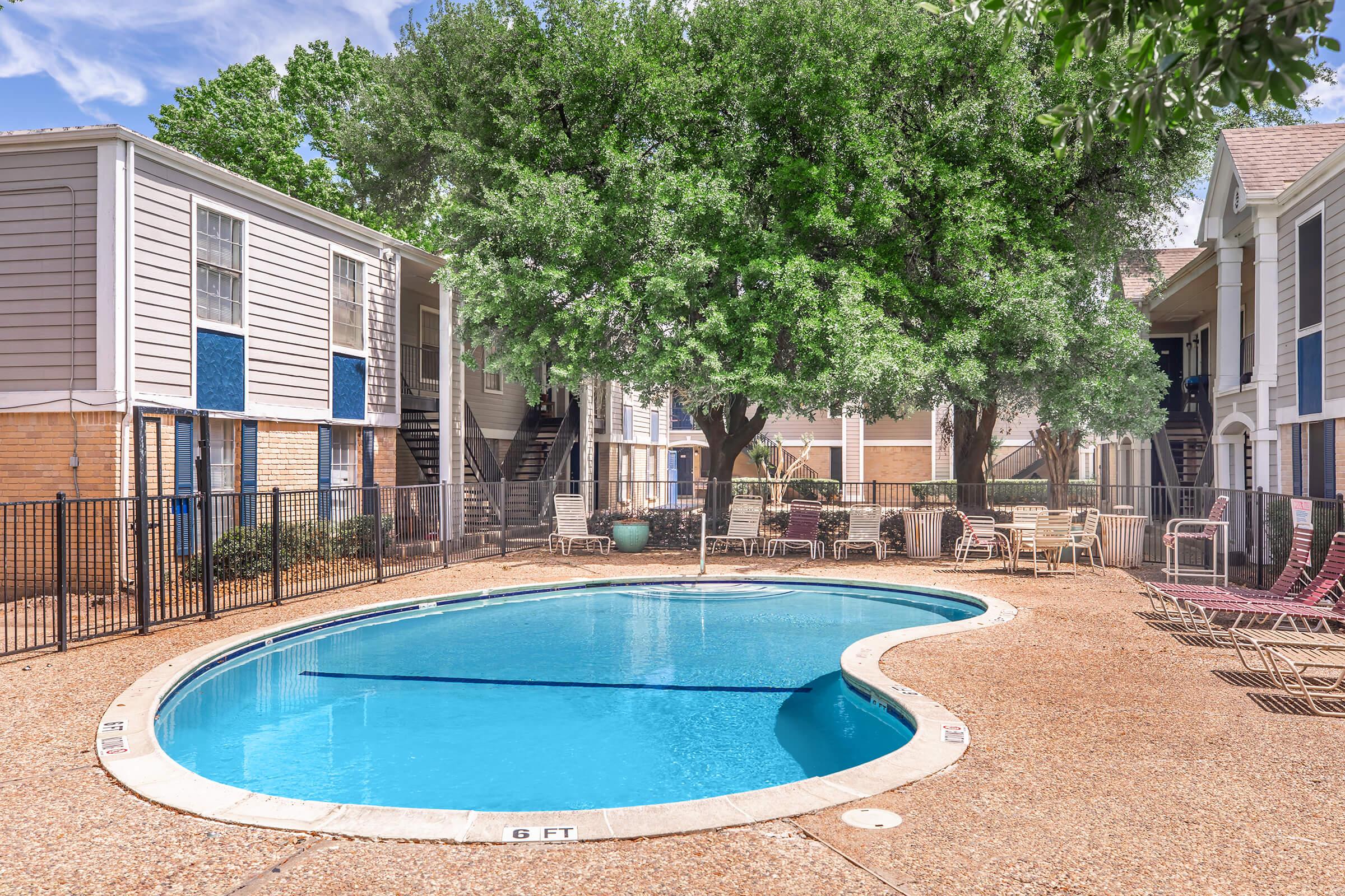 A rectangular swimming pool surrounded by a textured concrete deck, with lounge chairs and tables nearby. Lush green trees provide shade around the pool area, which is enclosed by a black fence and features two adjacent apartment buildings with blue accents on the windows. Clear blue sky visible overhead.