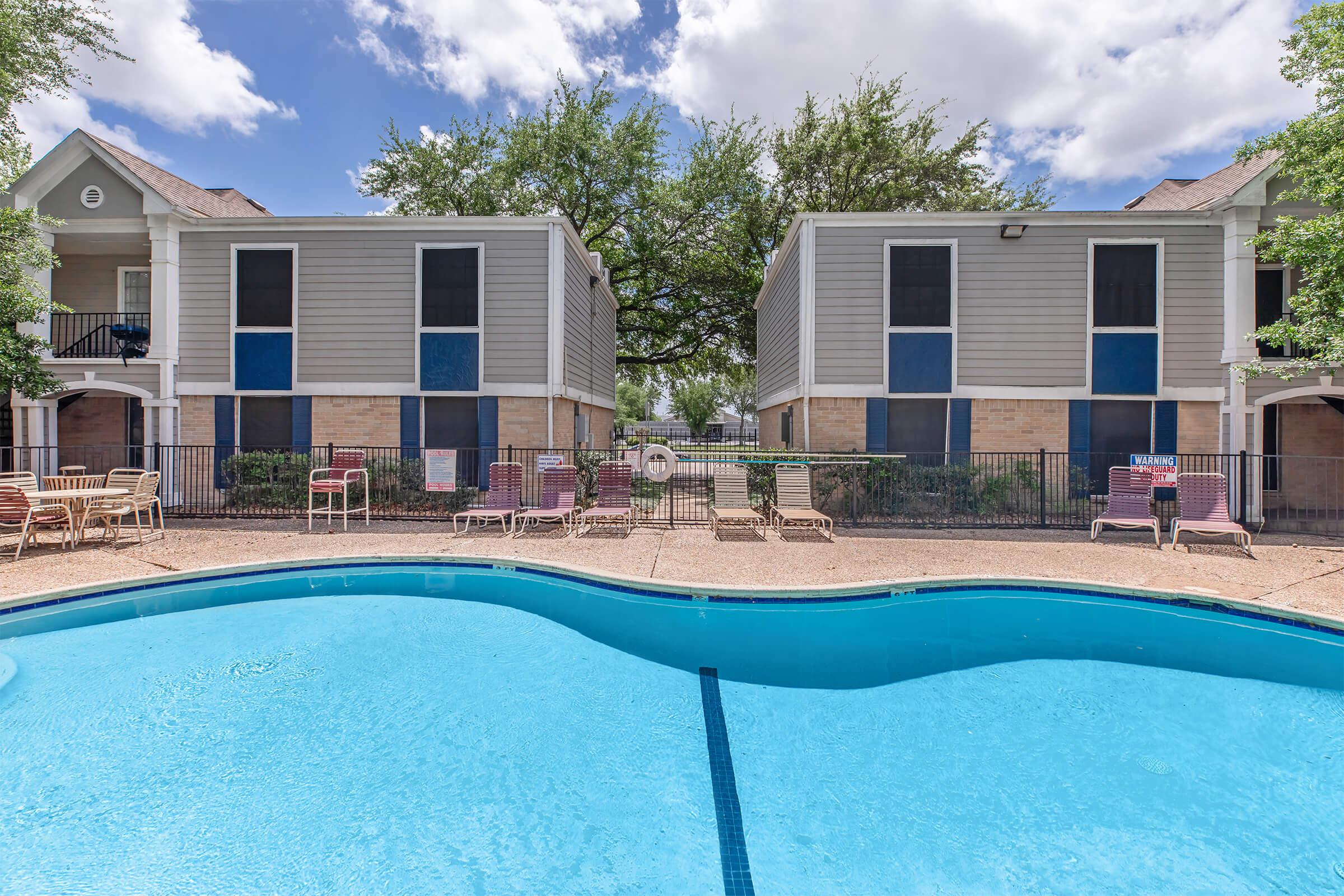 A swimming pool surrounded by lounge chairs is situated between two residential buildings. The pool features a curved shape, and the area is enclosed with a fence. Lush green trees provide shade in the background under a partly cloudy sky.