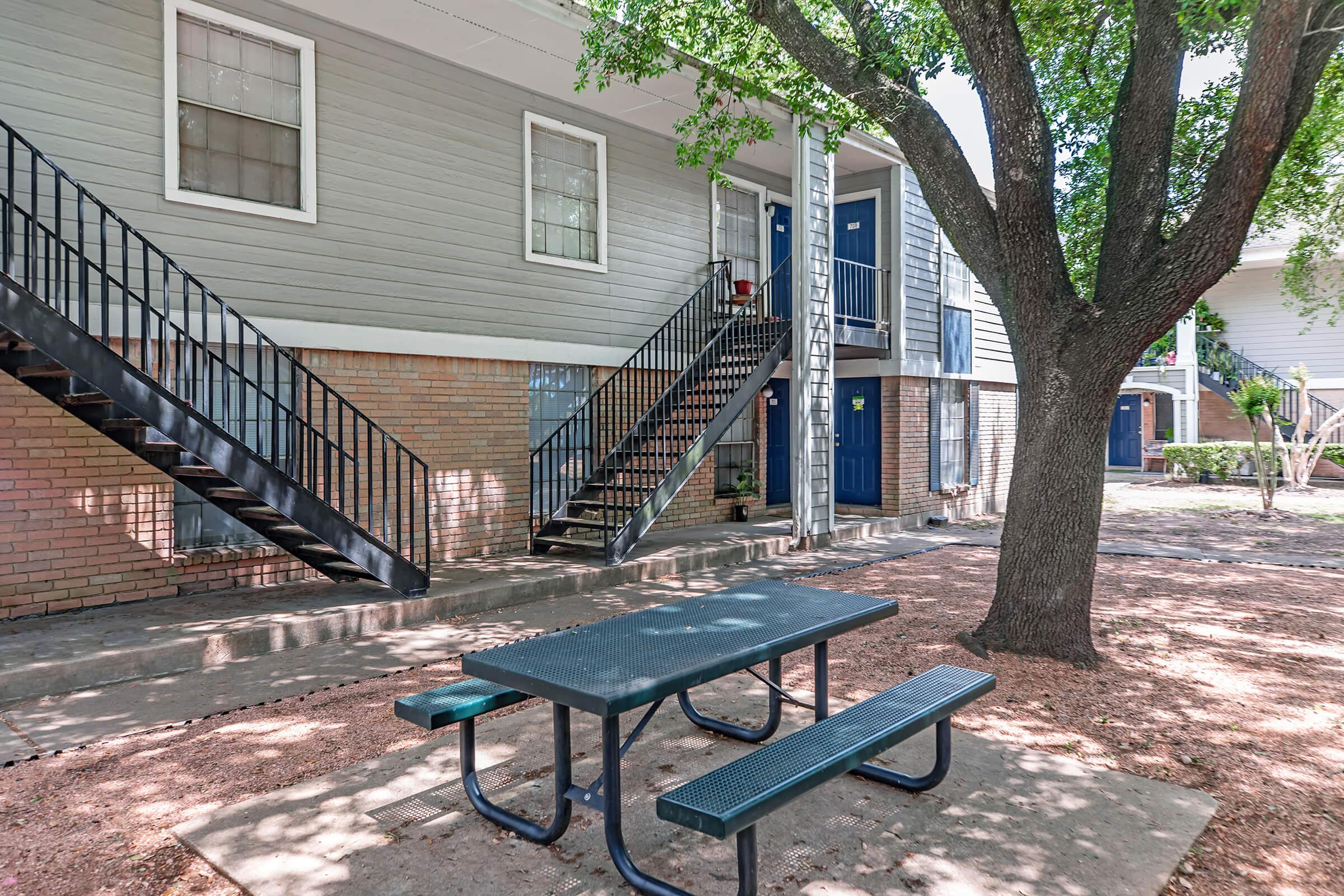 A view of a multi-story apartment building with a light gray exterior and brick accents. There are two sets of black metal stairs leading to entrances on the second floor. A picnic table with benches sits underneath a large tree in the foreground, surrounded by a gravel area and greenery.