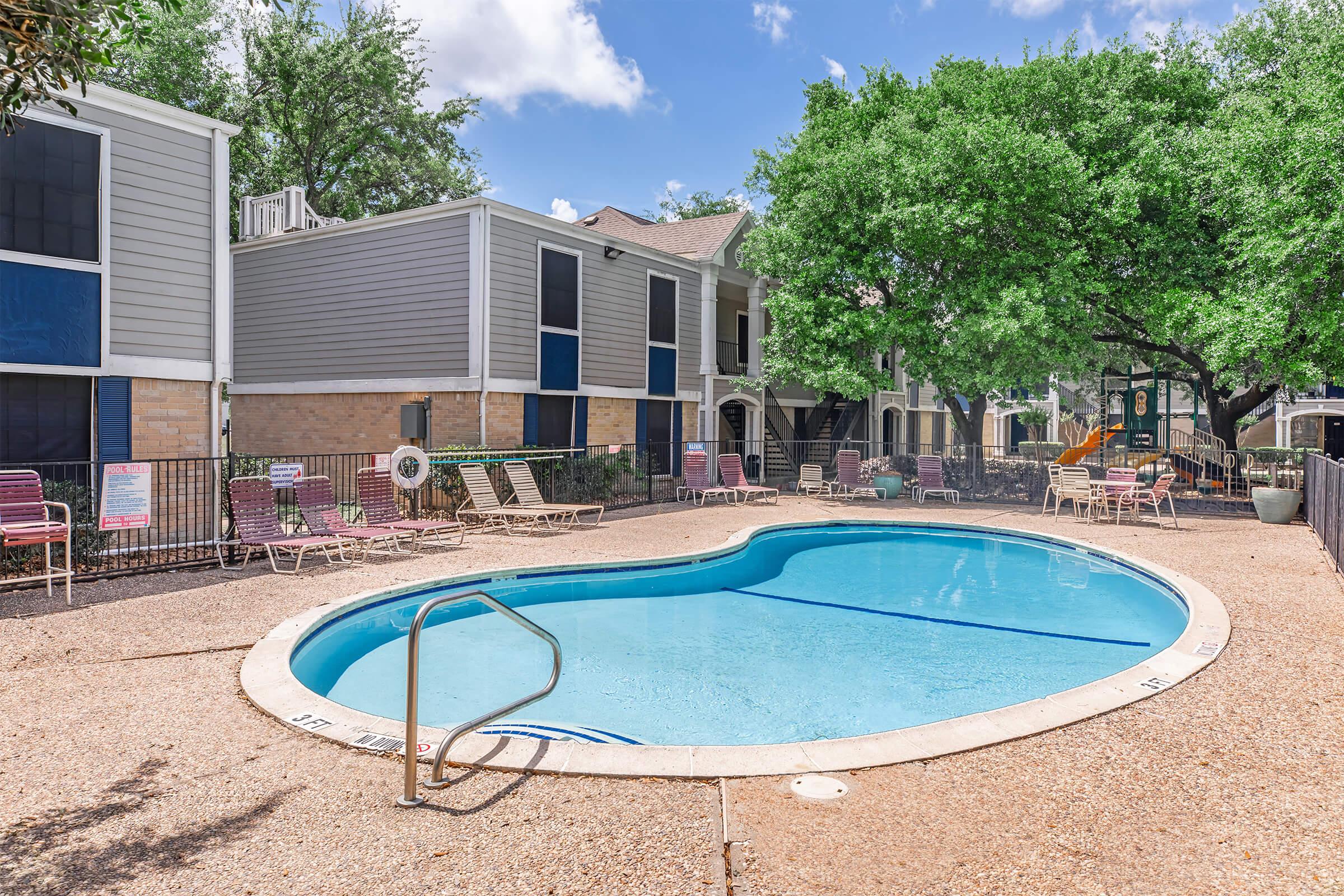 A clear swimming pool with a curved shape, surrounded by lounge chairs, located in a courtyard with green trees. Light gray and beige apartment buildings are in the background, with a blue sky and fluffy clouds above. The area appears inviting and well-maintained, perfect for relaxation.