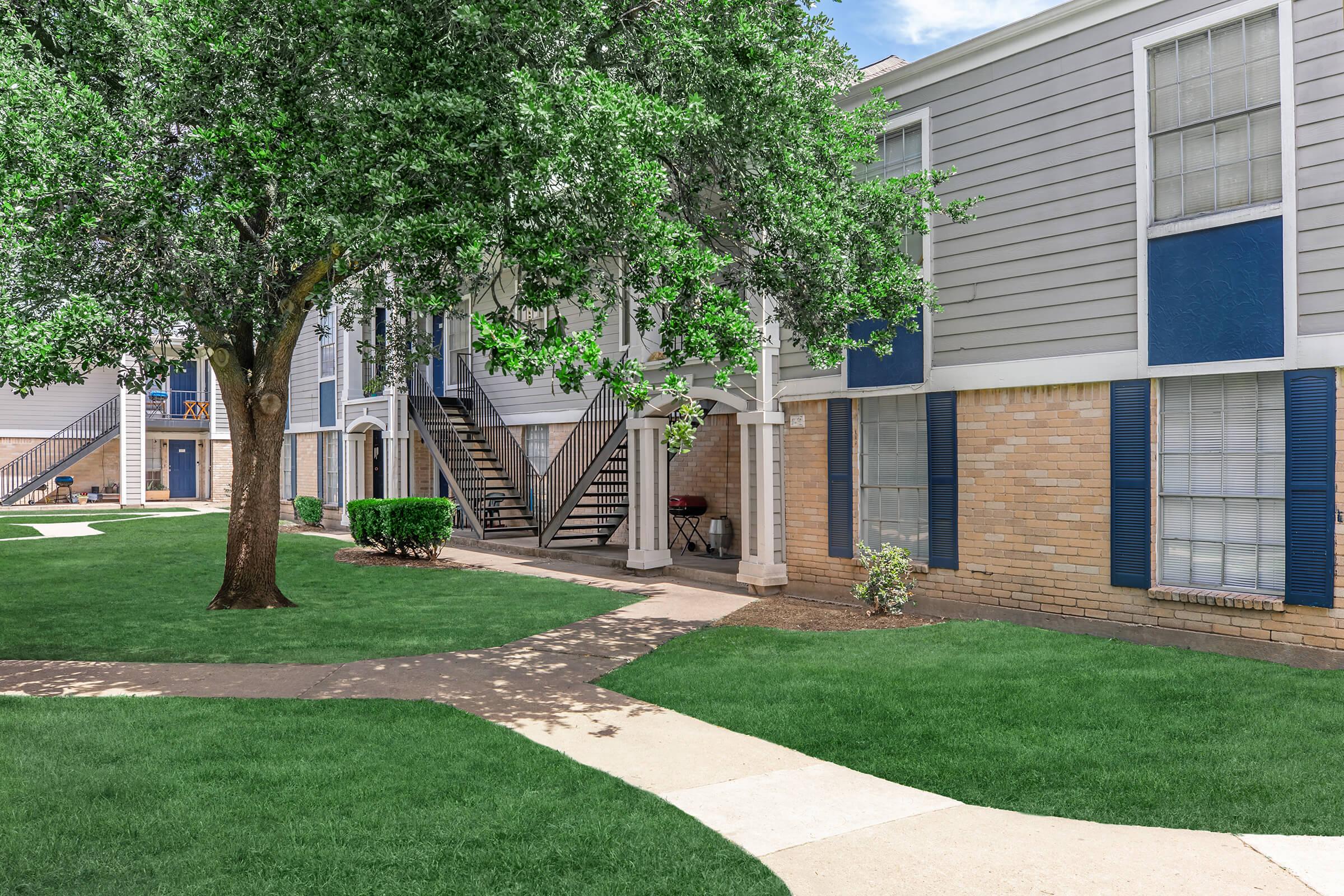 A landscaped residential complex featuring two-story buildings with gray siding and blue shutters. A large tree and neatly trimmed shrubs are present in the foreground, along with a walkway that leads through a green lawn. Stairs access the upper units, and there is a clear blue sky in the background.