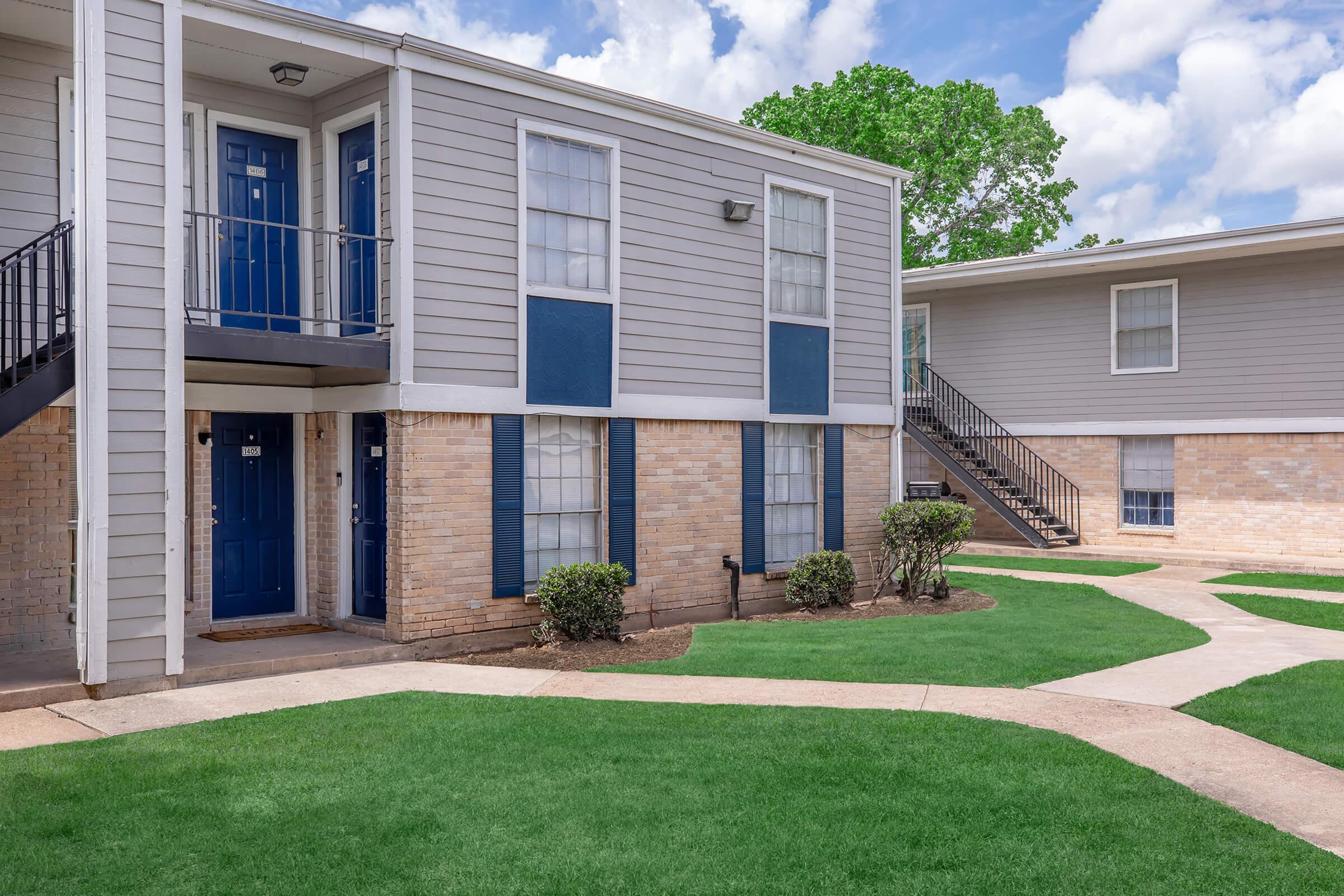 Exterior view of a two-story apartment building with blue doors and white trim. The building features brick accents and a well-maintained lawn with pathways. In the background, there are some trees and a partly cloudy sky.