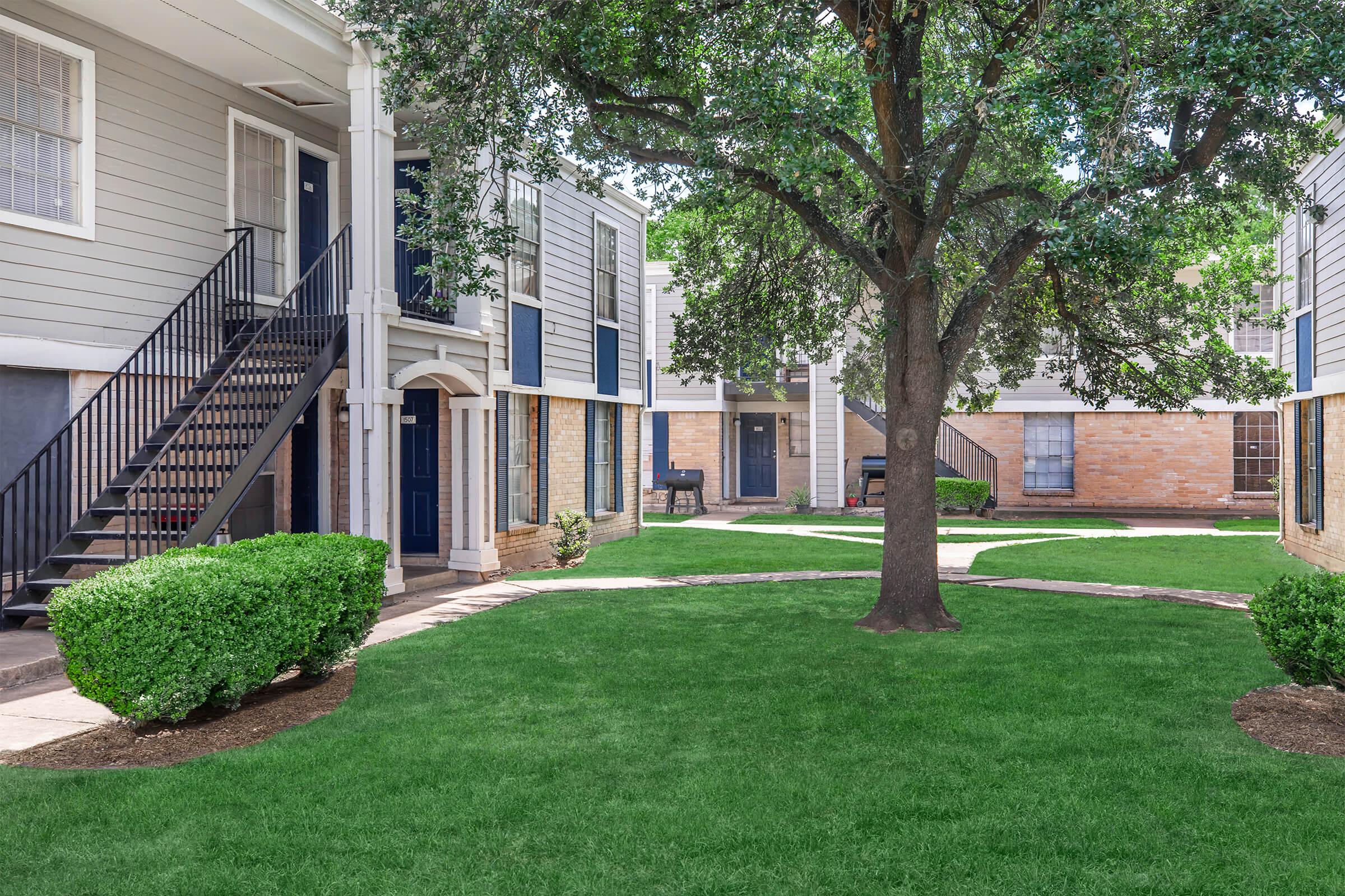 A landscaped courtyard featuring a large tree and manicured grass, surrounded by two-story apartment buildings. The buildings have blue and gray siding, with stairs leading to upper units. There are benches and seating areas visible in the background, creating a serene residential atmosphere.