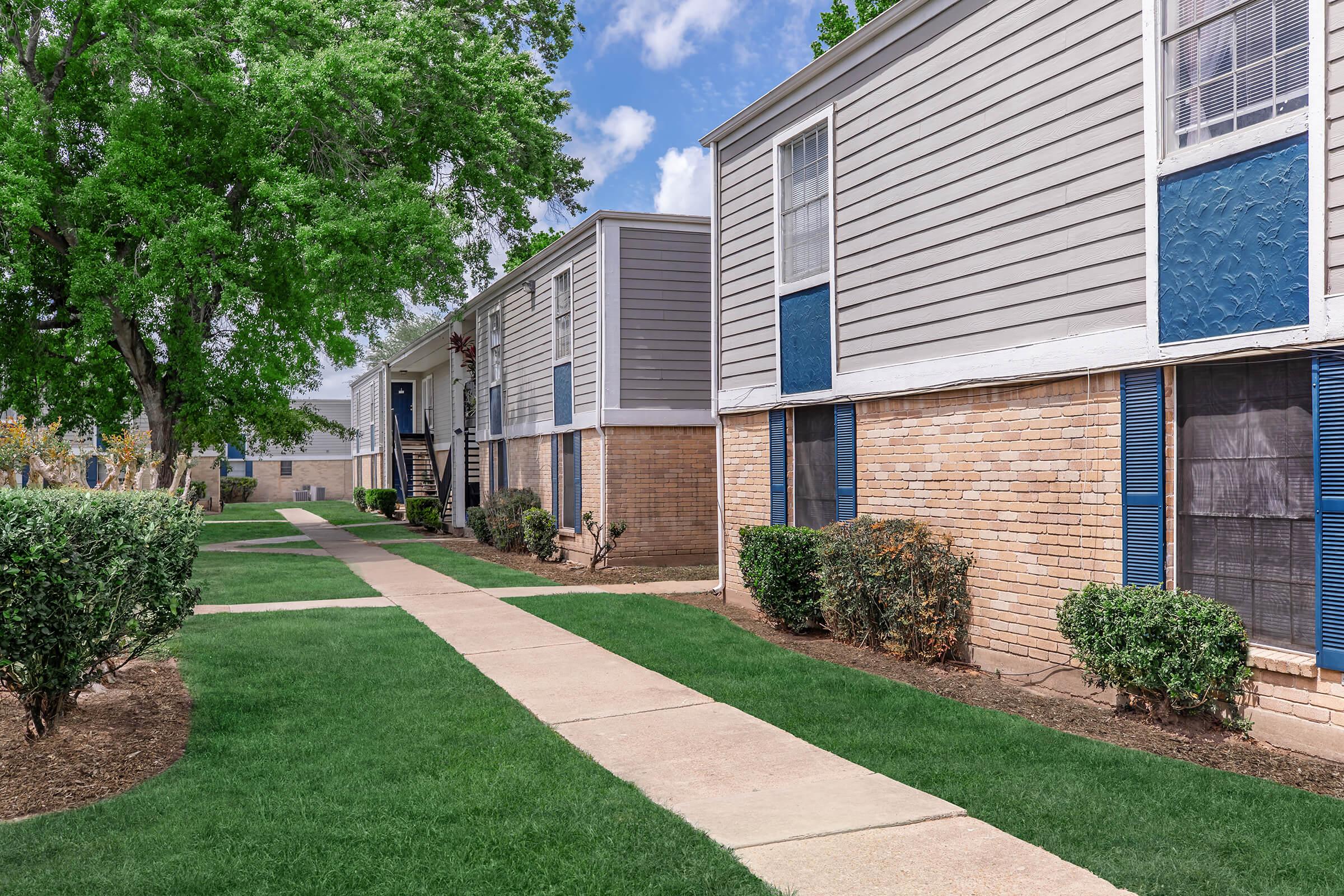 A well-maintained pathway lined with green grass leads to a row of apartment buildings. The buildings feature gray siding with blue accents and large windows. Small bushes are planted along the pathway, and there are trees in the background under a clear blue sky, creating a pleasant residential atmosphere.
