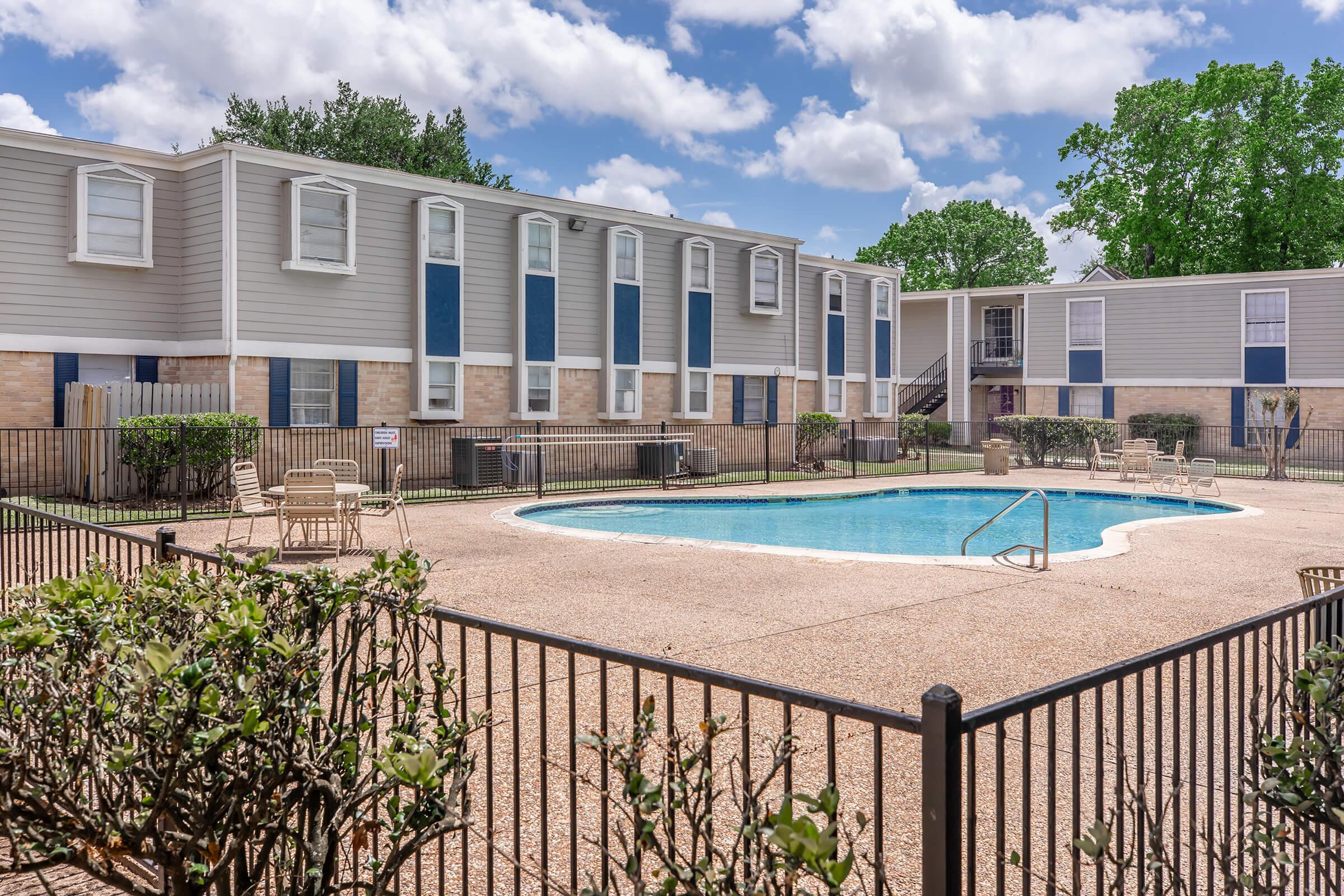A swimming pool surrounded by a fenced area, with lounge chairs positioned on a paved deck. In the background, two residential buildings with gray siding and blue windows are visible. The scene is set under a partly cloudy sky, and there are trees and shrubs adding greenery to the area.