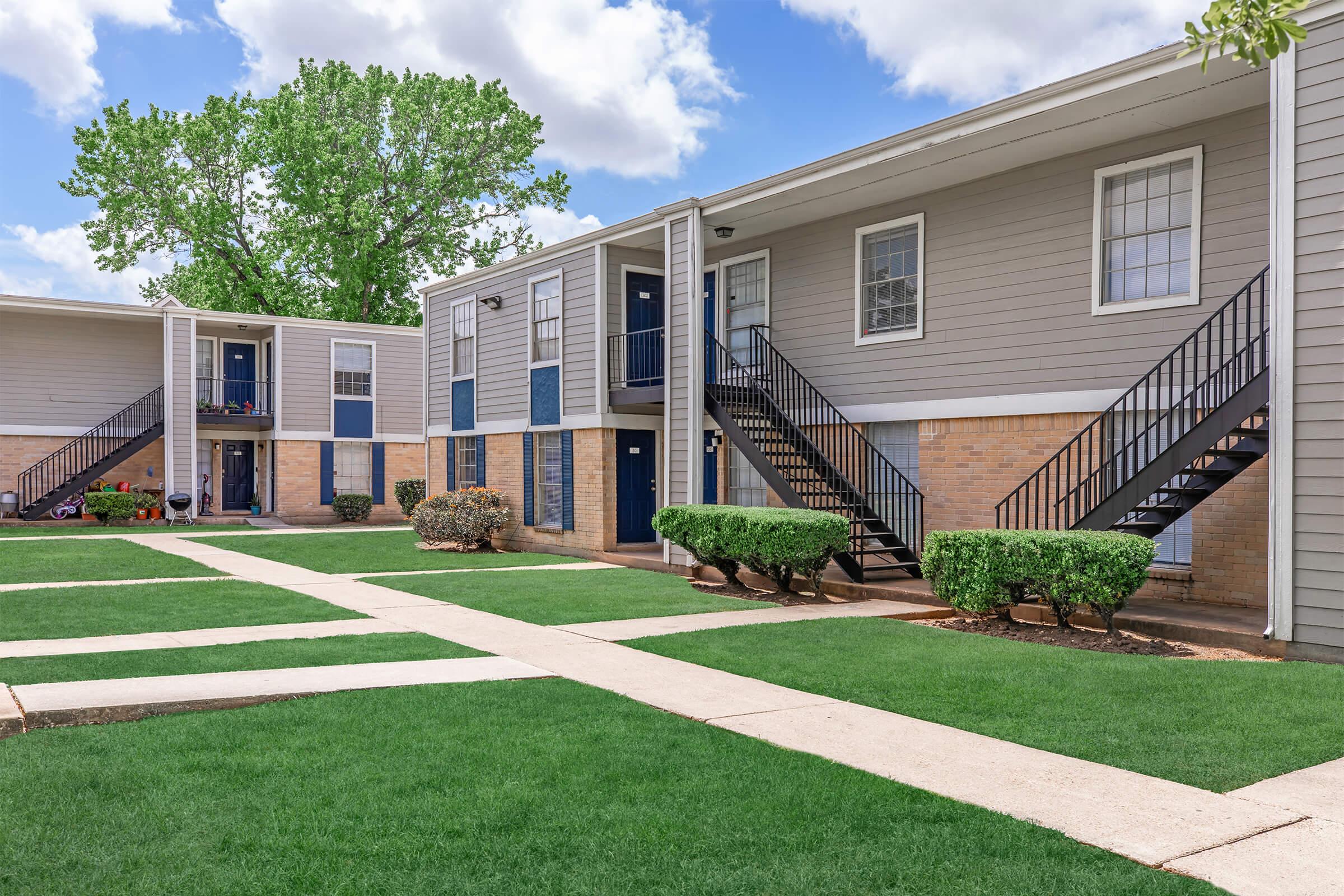 A well-maintained apartment complex featuring two-story buildings with blue doors and black staircases. The green lawn is neatly trimmed, and there are shrubs along the walkways. The sky is partly cloudy, adding to the pleasant outdoor atmosphere.