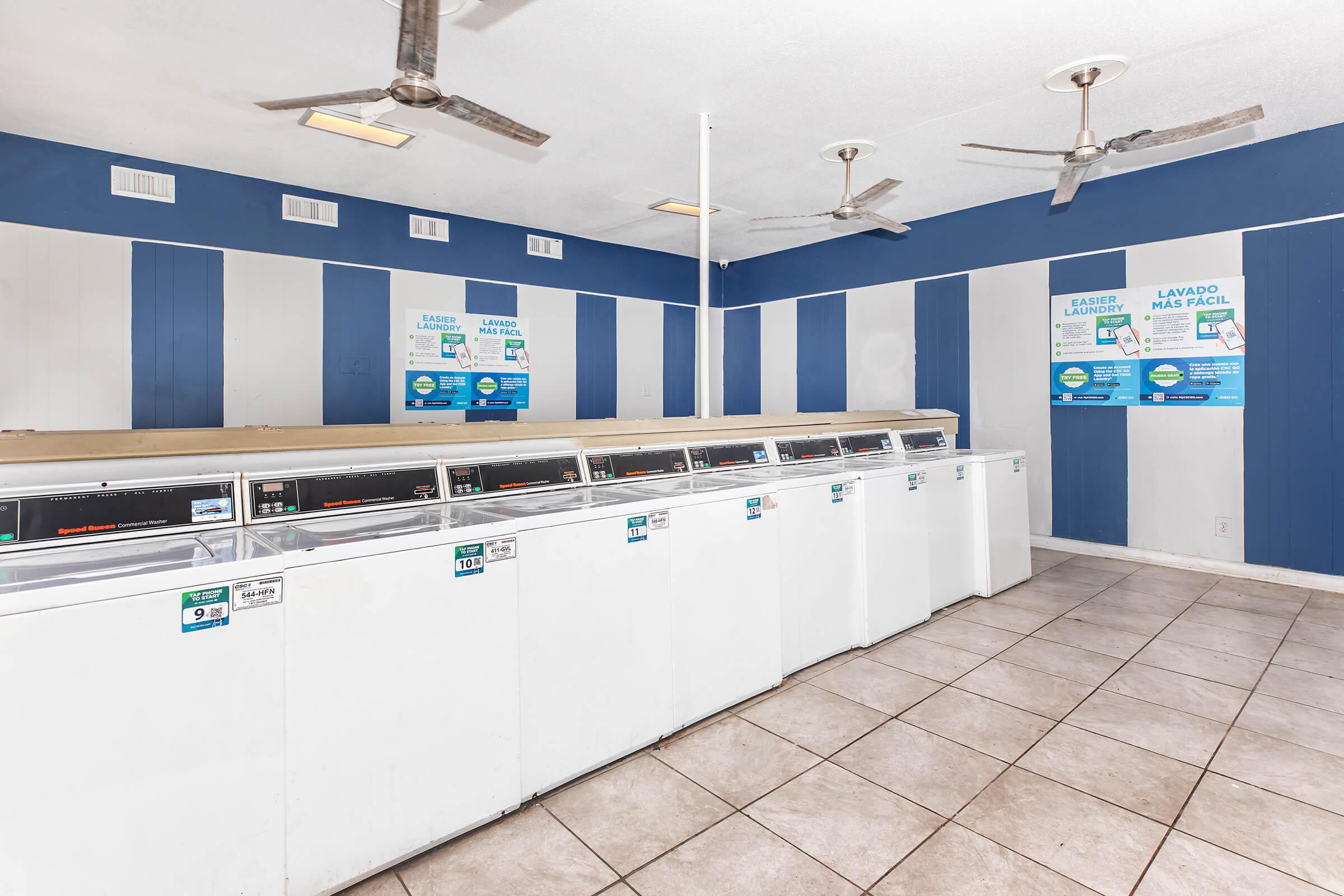 A clean and well-lit laundromat featuring rows of white washing machines against a blue and gray striped wall. Ceiling fans provide ventilation. Posters with laundry instructions and payment options are displayed on the wall. The floor is tiled, and the space is organized and inviting.