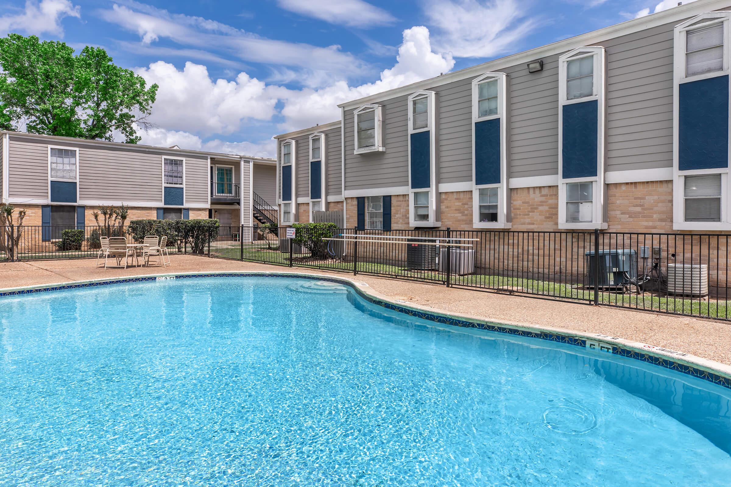 A bright, inviting swimming pool surrounded by a fenced area, with lounge chairs placed nearby. In the background, two multi-story apartment buildings with blue and gray exteriors are visible under a partly cloudy sky. Lush greenery is present, enhancing the outdoor atmosphere.