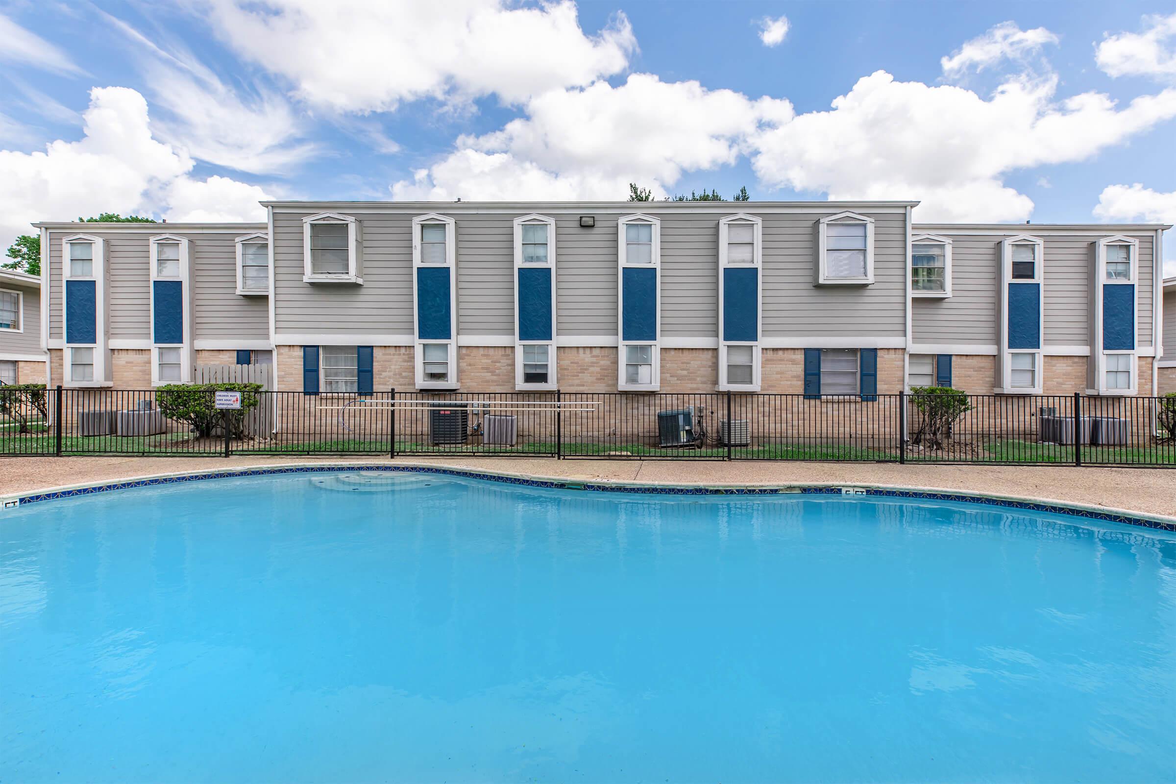 A clear blue swimming pool in the foreground, with a fenced area. In the background, a two-story residential building featuring multiple windows and a light gray exterior. The sky above is partly cloudy, creating a bright and inviting atmosphere.