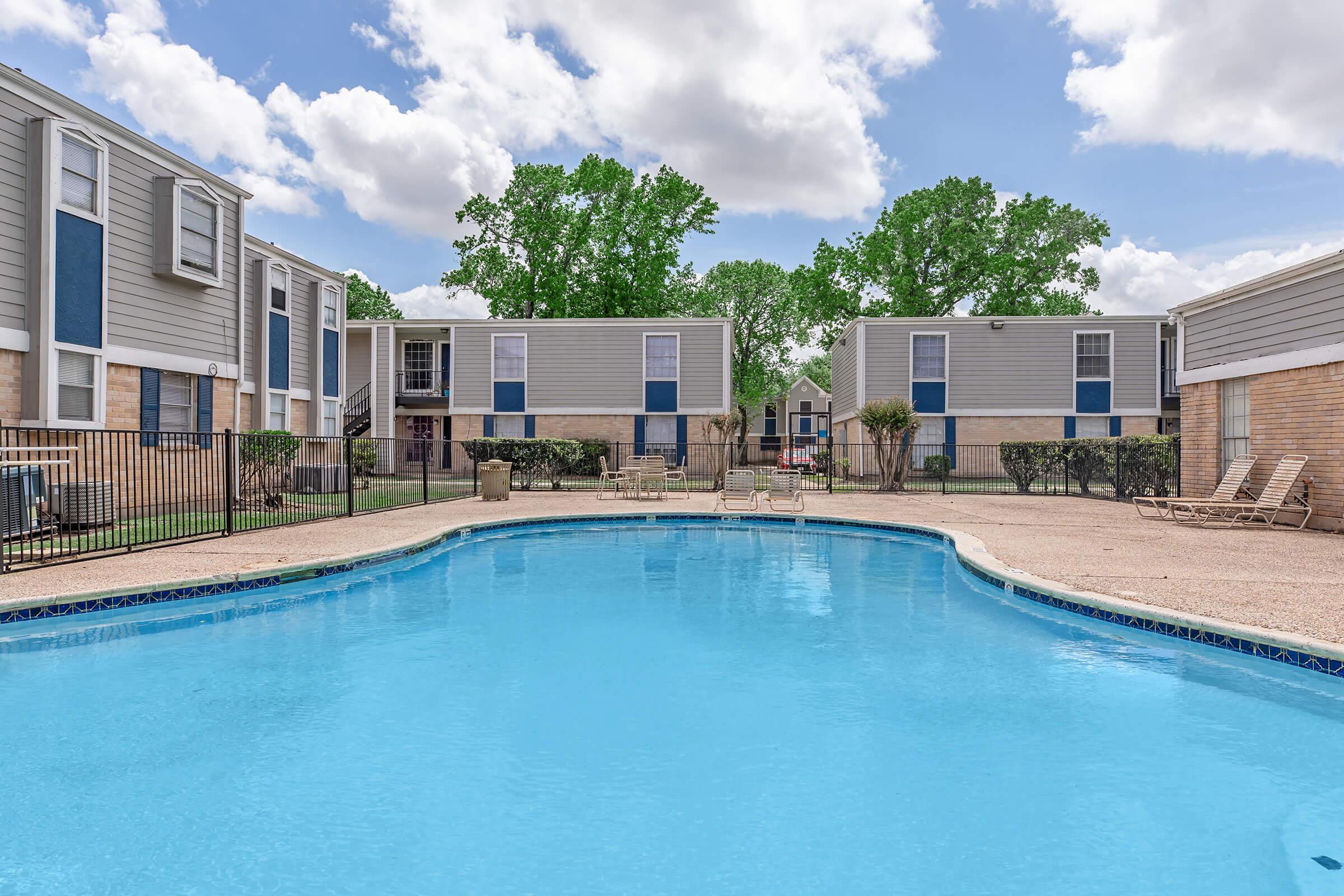 A bright blue swimming pool surrounded by a fence, with lounge chairs positioned on the deck. In the background, apartment buildings with gray siding and green trees under a partly cloudy sky. The scene conveys a serene and inviting outdoor space for relaxation.